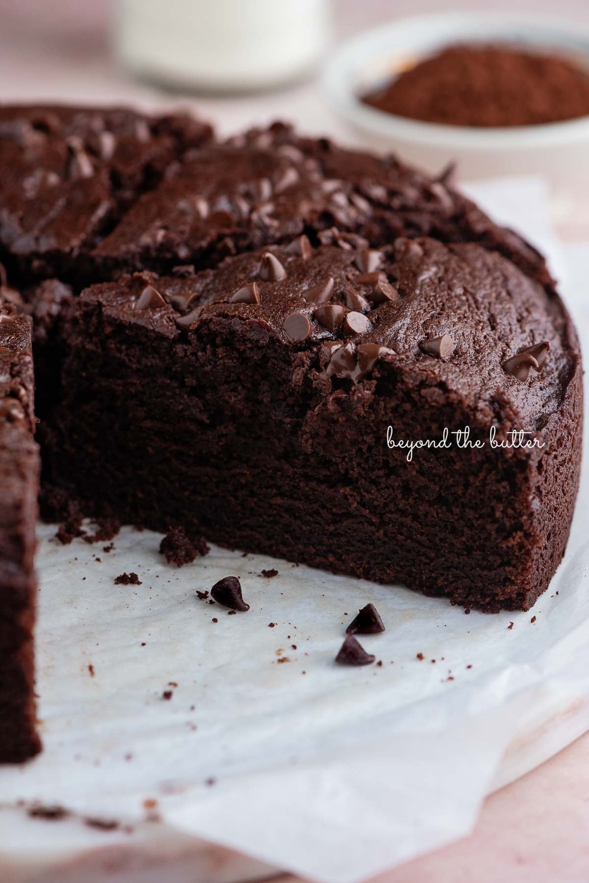 Slices of chocolate ricotta cake on white parchment paper and a white marble cake plate placed on a pink tiled background with a glass of milk and small bowl of cocoa powder nearby.