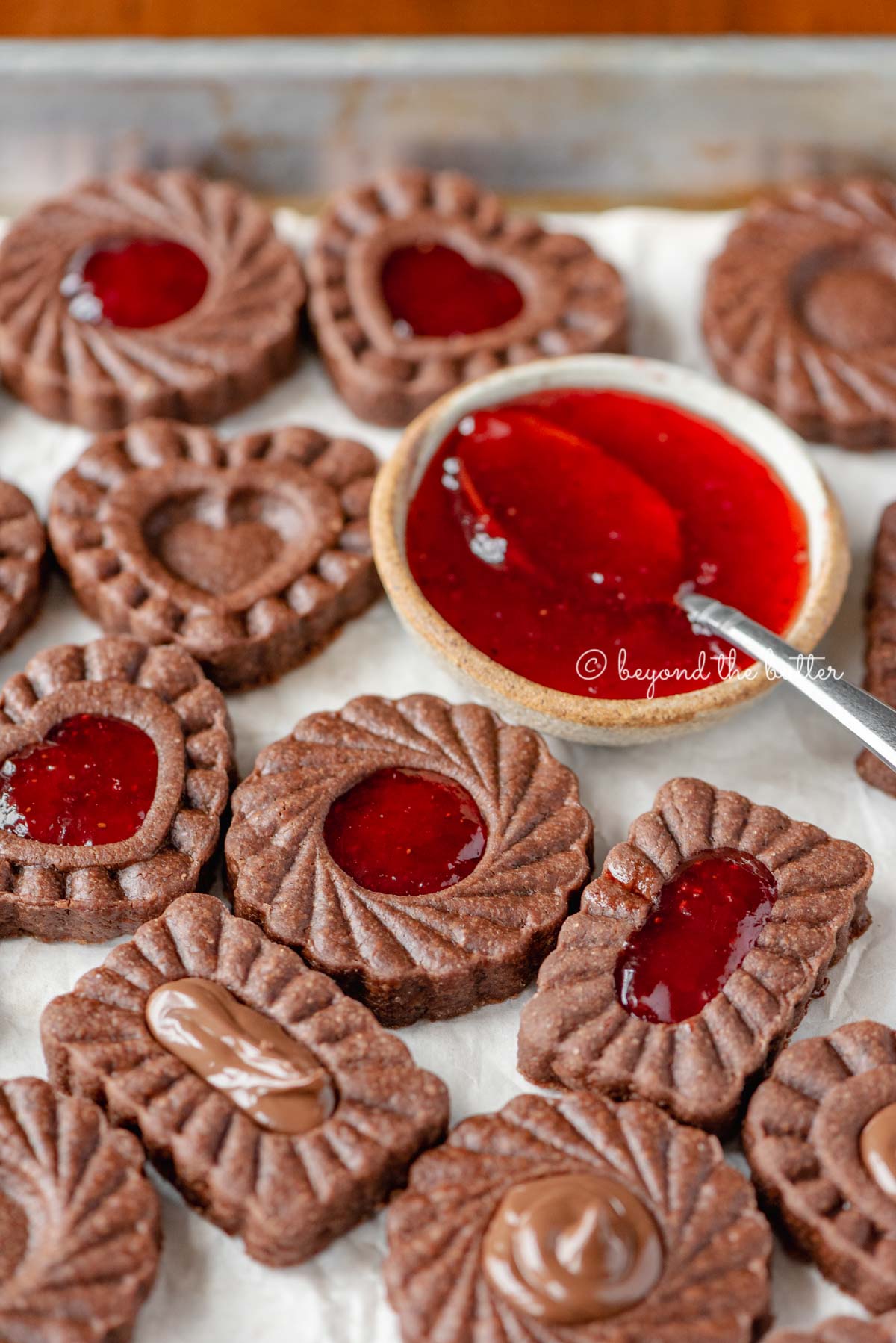 Parchment lined baking sheet full of chocolate thumbprint cookies with small bowl of strawberry jam and spoon | All Images © Luyiz®