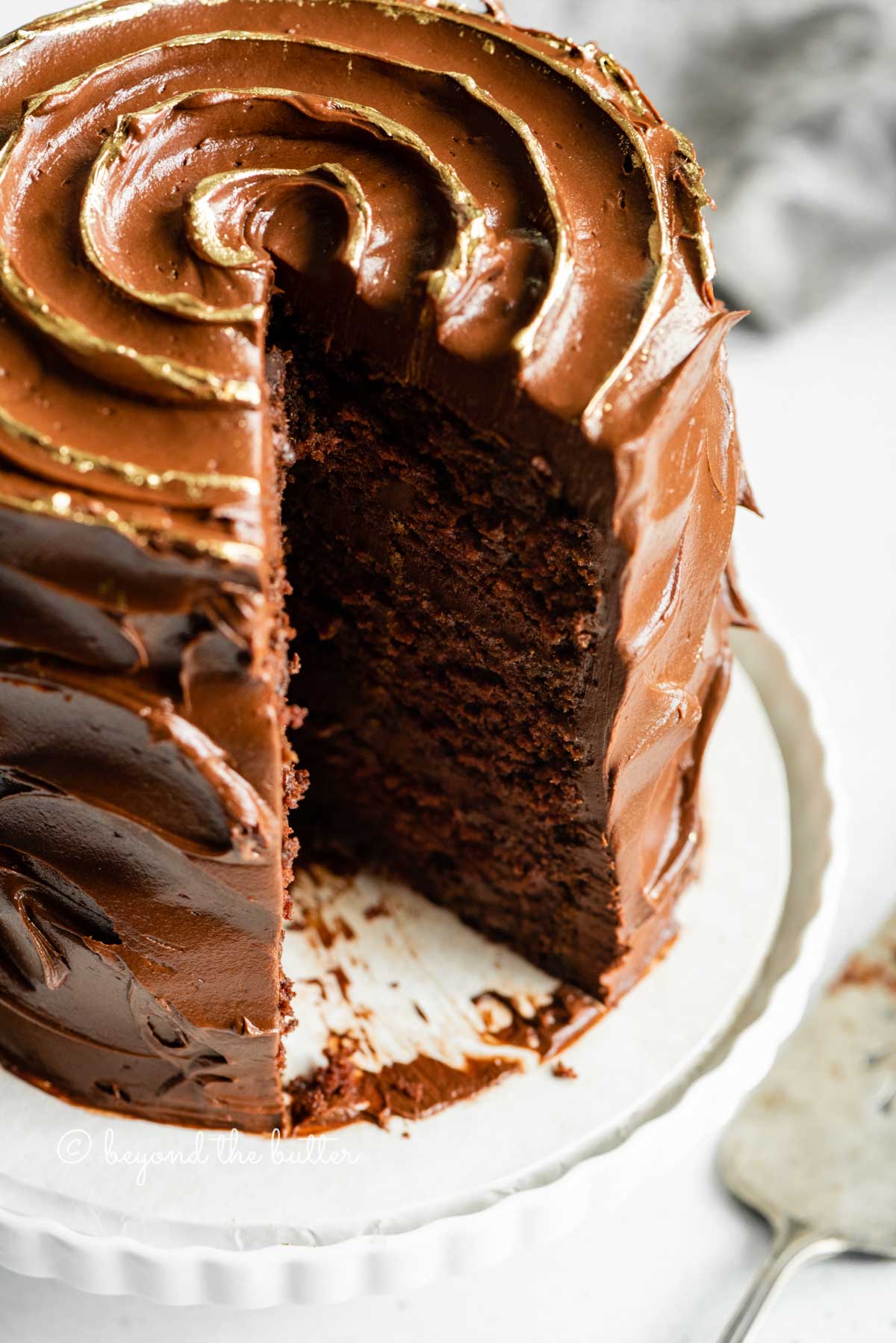 Angled closeup image of chocolate zucchini cake with gold decorative swirl on top and slice removed | All Images © Luyiz™