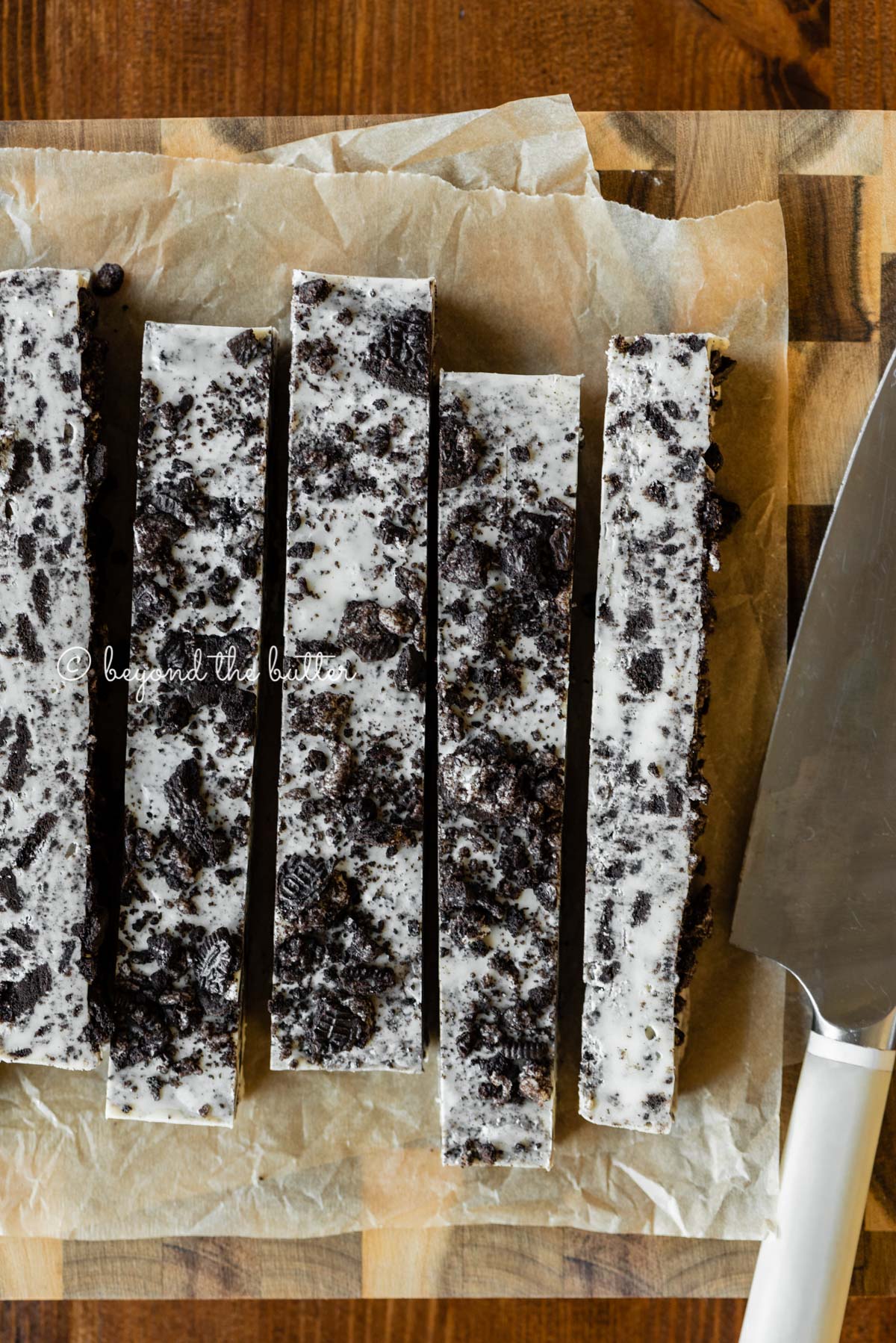 Bars of cookies and cream fudge on brown parchment paper and wood cutting board with knife on top | All Images © Luyiz®