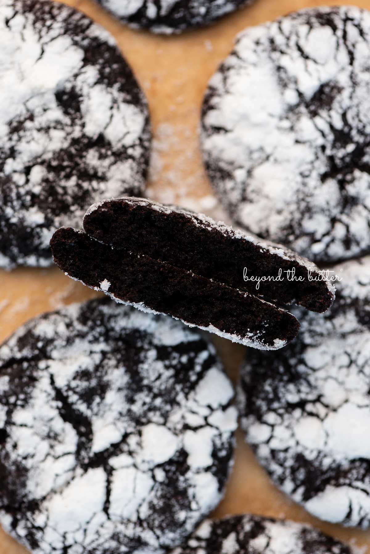 Split dark chocolate crinkle cookie to show the fudgy inside on parchment paper lined baking sheet | © Luyiz®