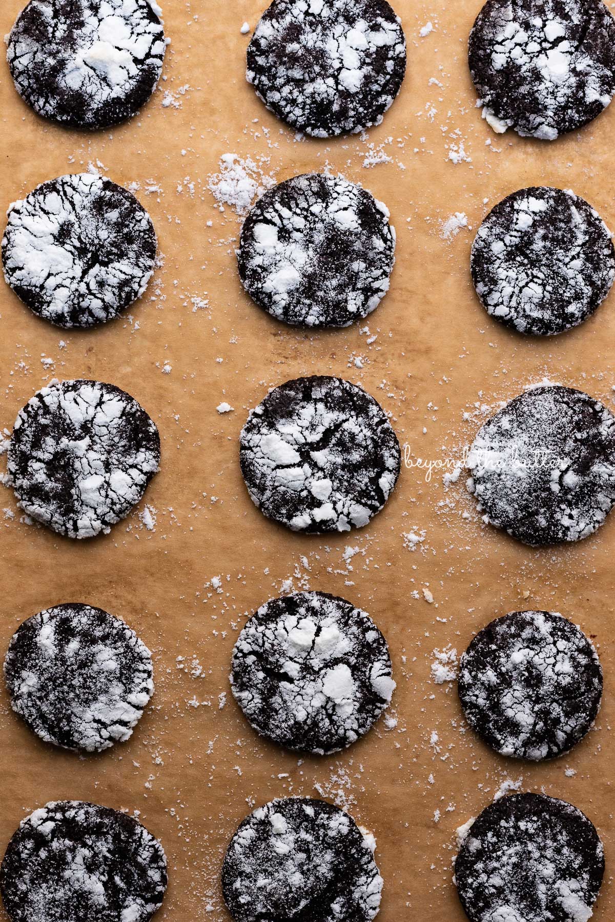 Dark chocolate crinkle cookies evenly spaced on a brown parchment paper lined baking sheet | © Luyiz®