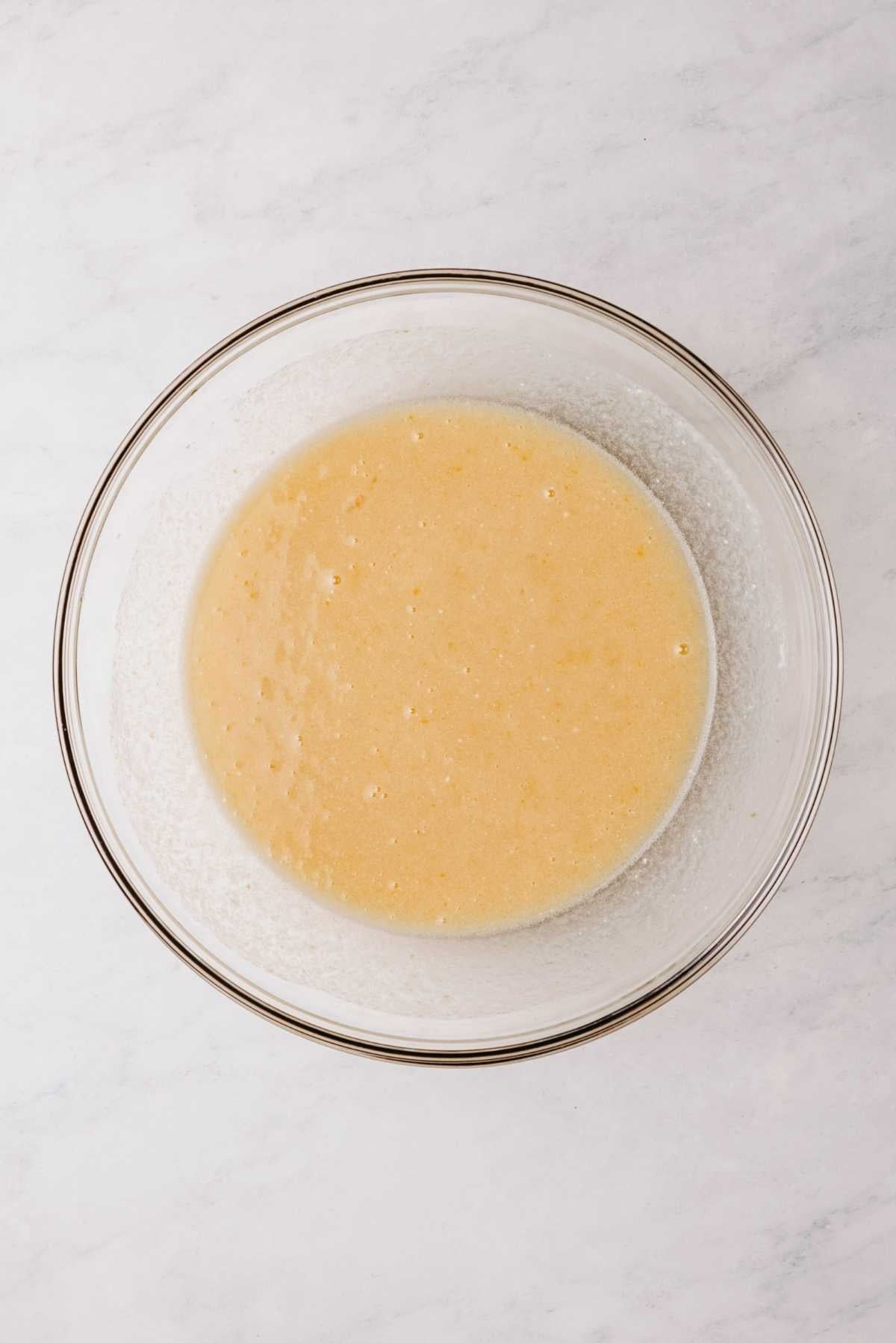 Whisked eggs, sugar, vegetable oil, vanilla extract, and buttermilk in a large glass mixing bowl on a white marbled background.