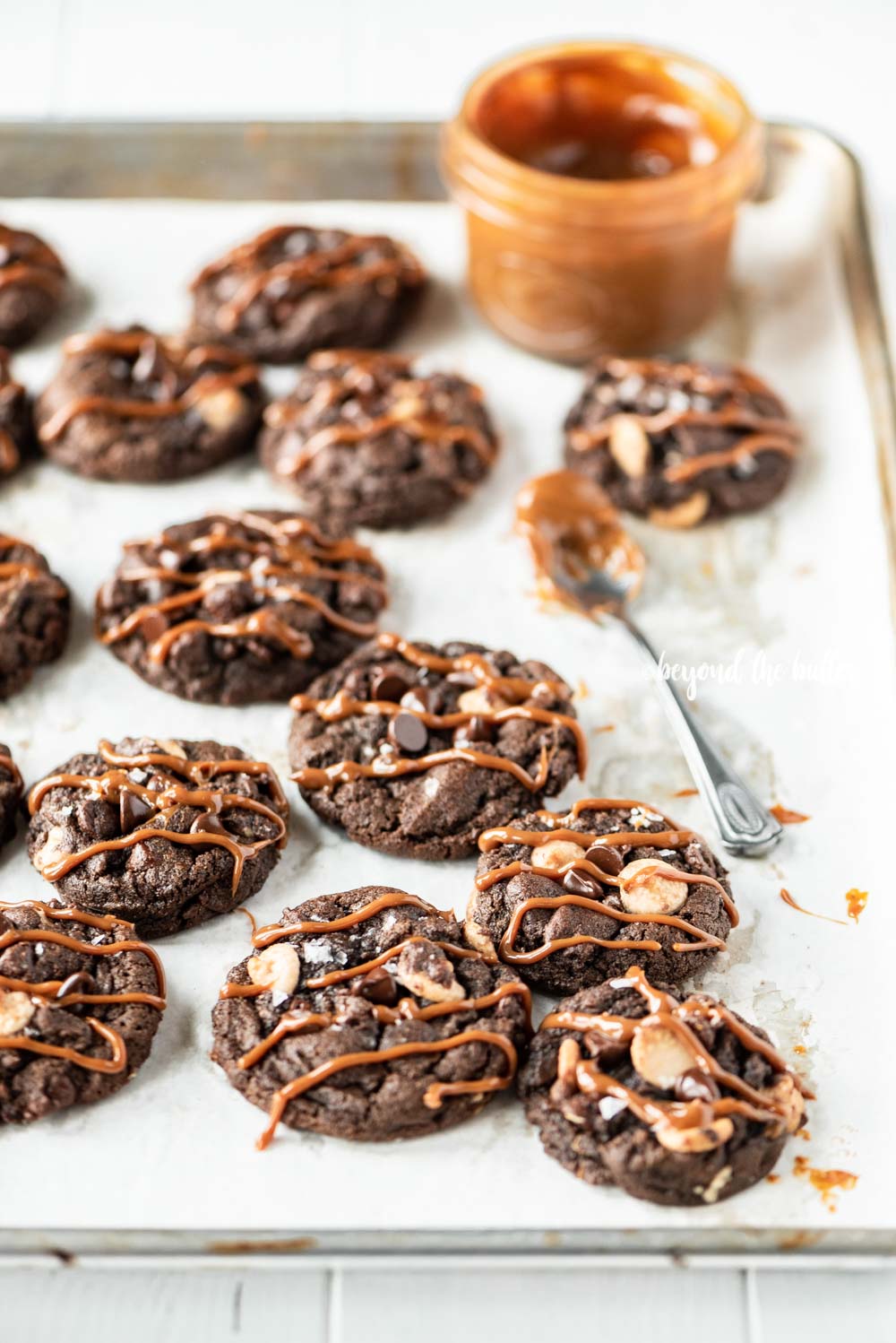 Overhead image of just baked double chocolate salted caramel cookies on a baking sheet with a spoon and jar of a caramel sauce resting close by | All Images © Luyiz™