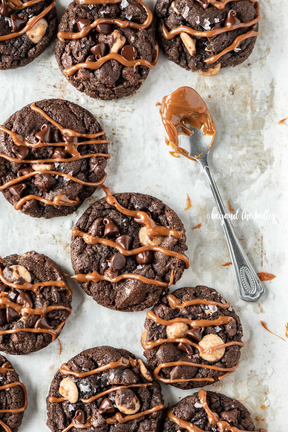 Overhead image of just baked double chocolate salted caramel cookies on a baking sheet with spoon dipped in a caramel sauce resting close by | All Images © Luyiz™
