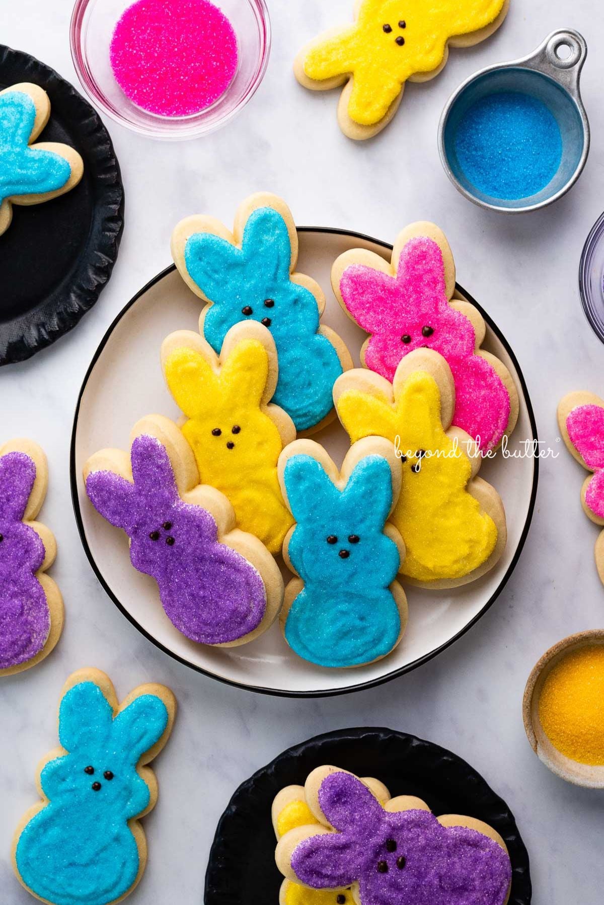 Plate full of Easter bunny cut out sugar cookies decorated in purple, pink, blue, and yellow sanding sugars with chocolate dots for eyes and noses on white marbled background.