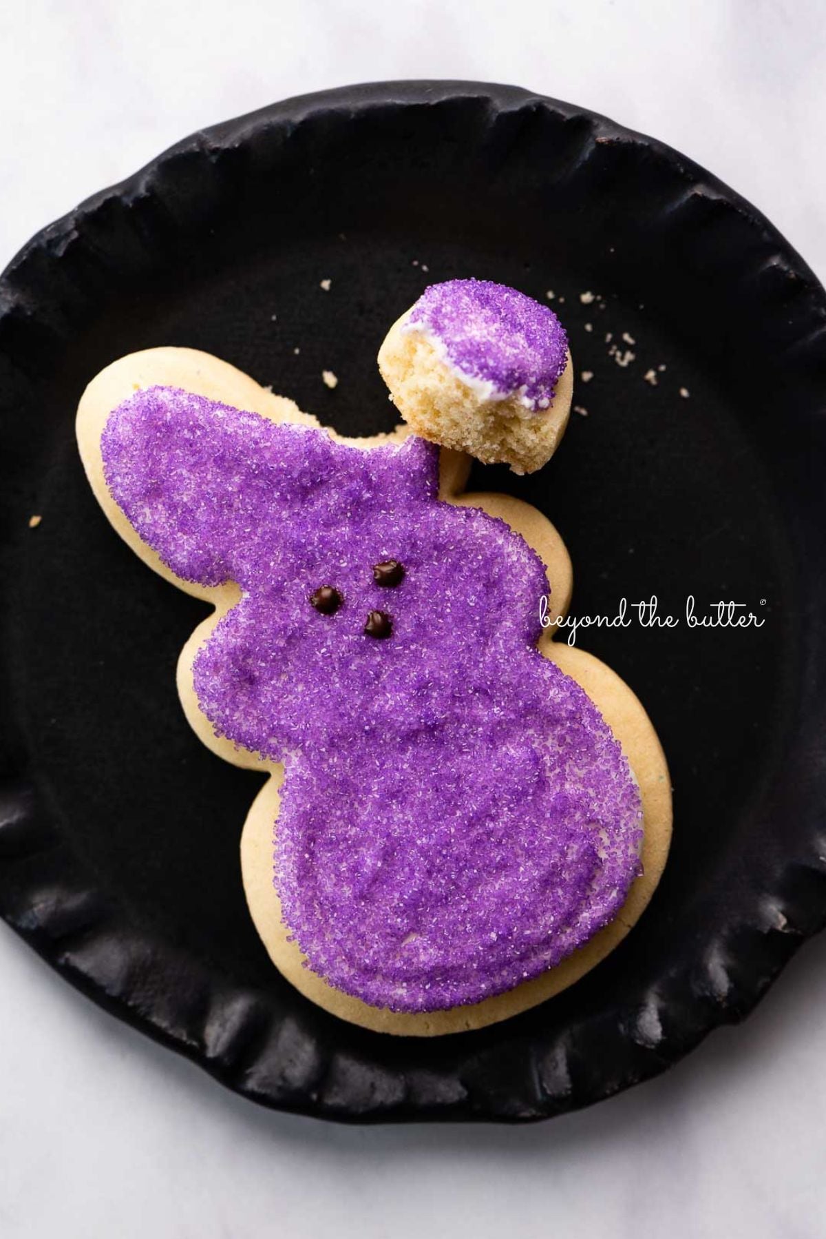 Easter bunny cut out sugar cookie decorated with purple sanding sugar and a chocolate nose and eyes with its right ear broken off on a black dessert plate.