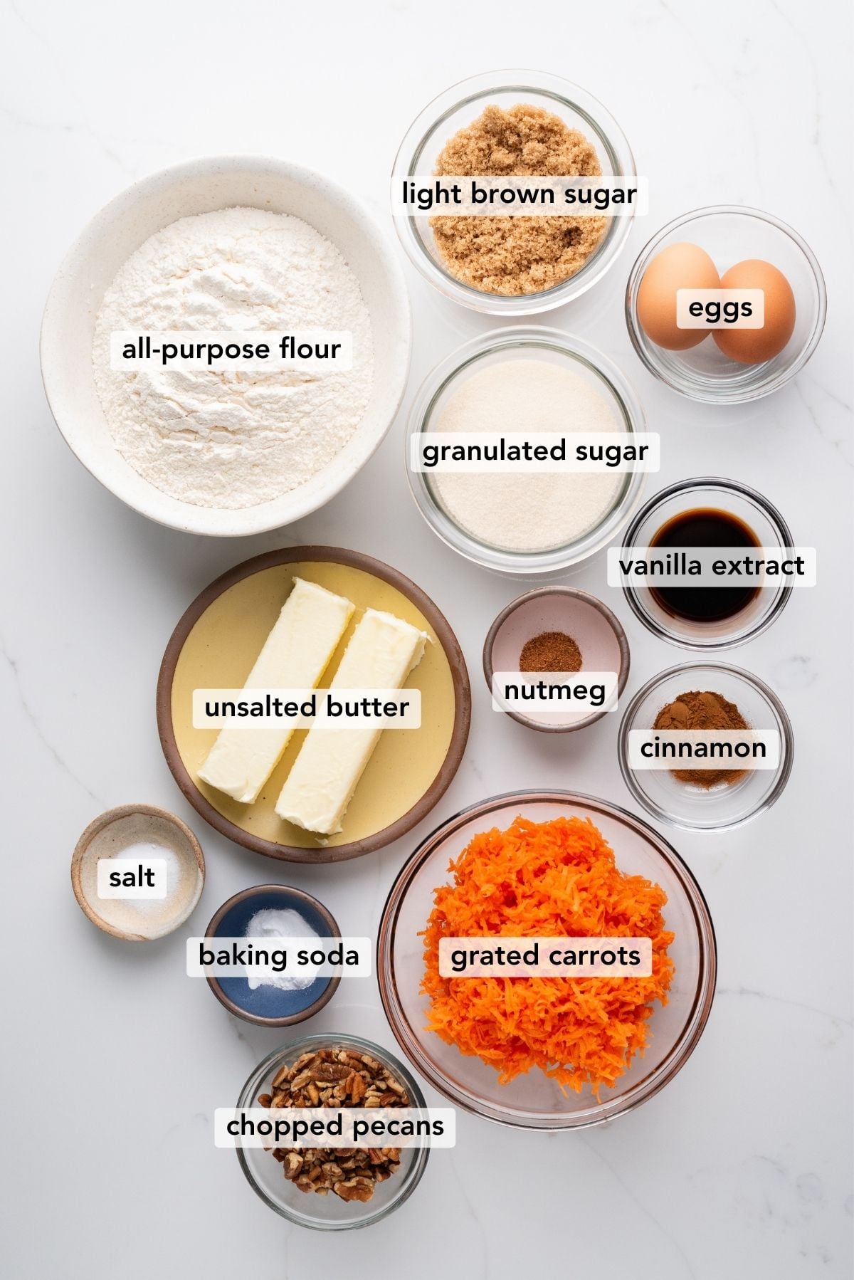 Carrot cake cookies ingredients in various size bowls labeled as all-purpose flour, baking soda, cinnamon, nutmeg, salt, unsalted butter, light brown sugar, granulated sugar, eggs, vanilla extract, grated carrots, and chopped pecans on a white marbled background.