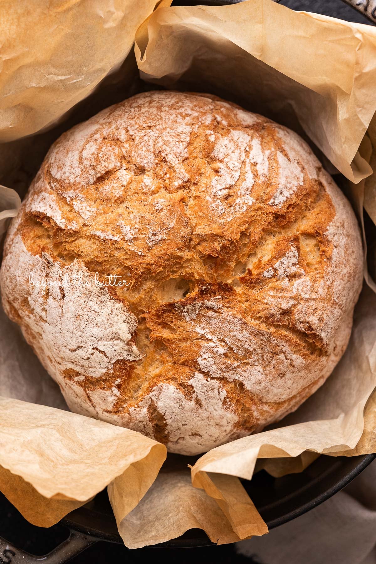Round loaf of french bread inside a dark gray dutch oven lined with a sheet of brown parchment paper on a black background.
