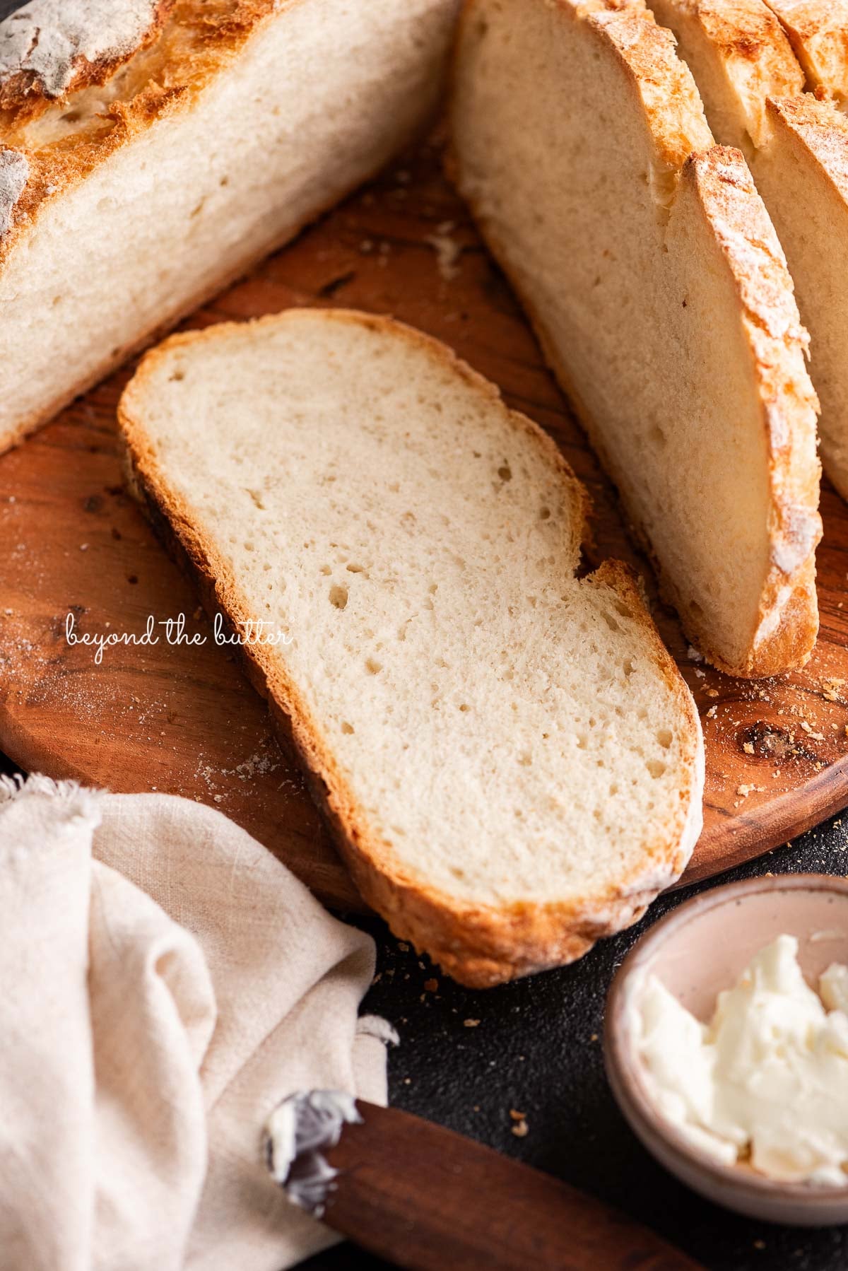Sliced french bread placed on a round wooden cutting board next to a small pink bowl of butter and a light beige cloth napkin on a black background.