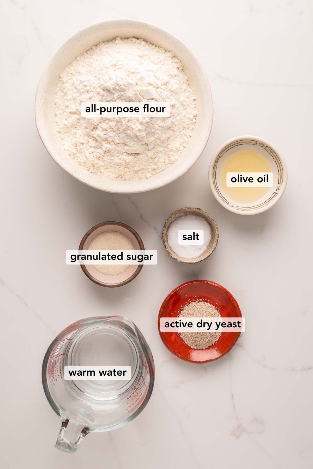 Ingredients in various size bowls labeled as all-purpose flour, olive oil, granulated sugar, salt, warm water, and active dry yeast on a white marbled background.