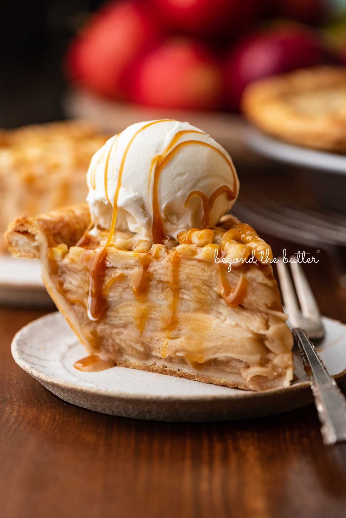 Slice of apple pie drizzled with caramel and topped with a scoop of vanilla bean ice cream on a ceramic dessert plate placed next to apple pie and bowl of apples on a wood background.