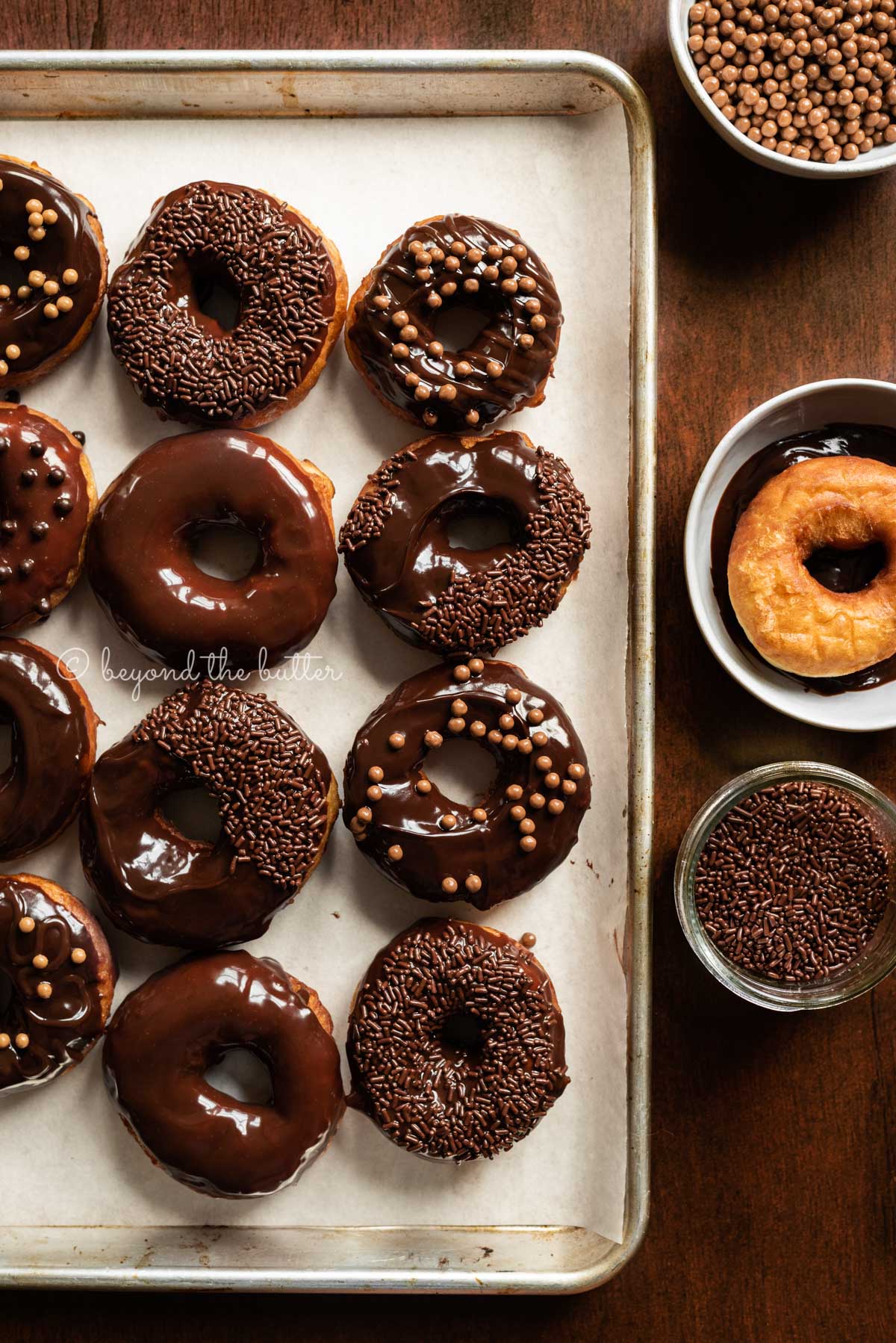 Chocolate glazed decorated donuts on a parchment paper lined baking sheet with a donut and chocolate jimmies next to them | All Images © Luyiz™