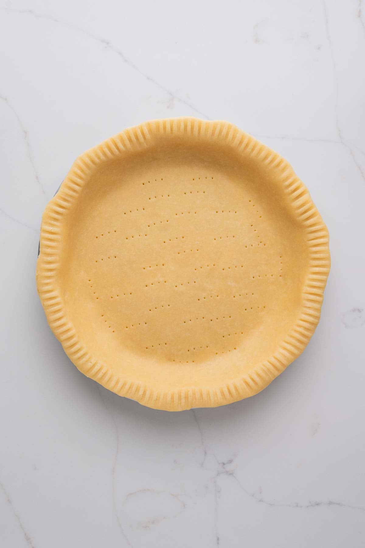 Rolled out pie dough placed in a metal pie tin that's been trimmed, edges crimped and bottom docked with a fork on a white marbled background.