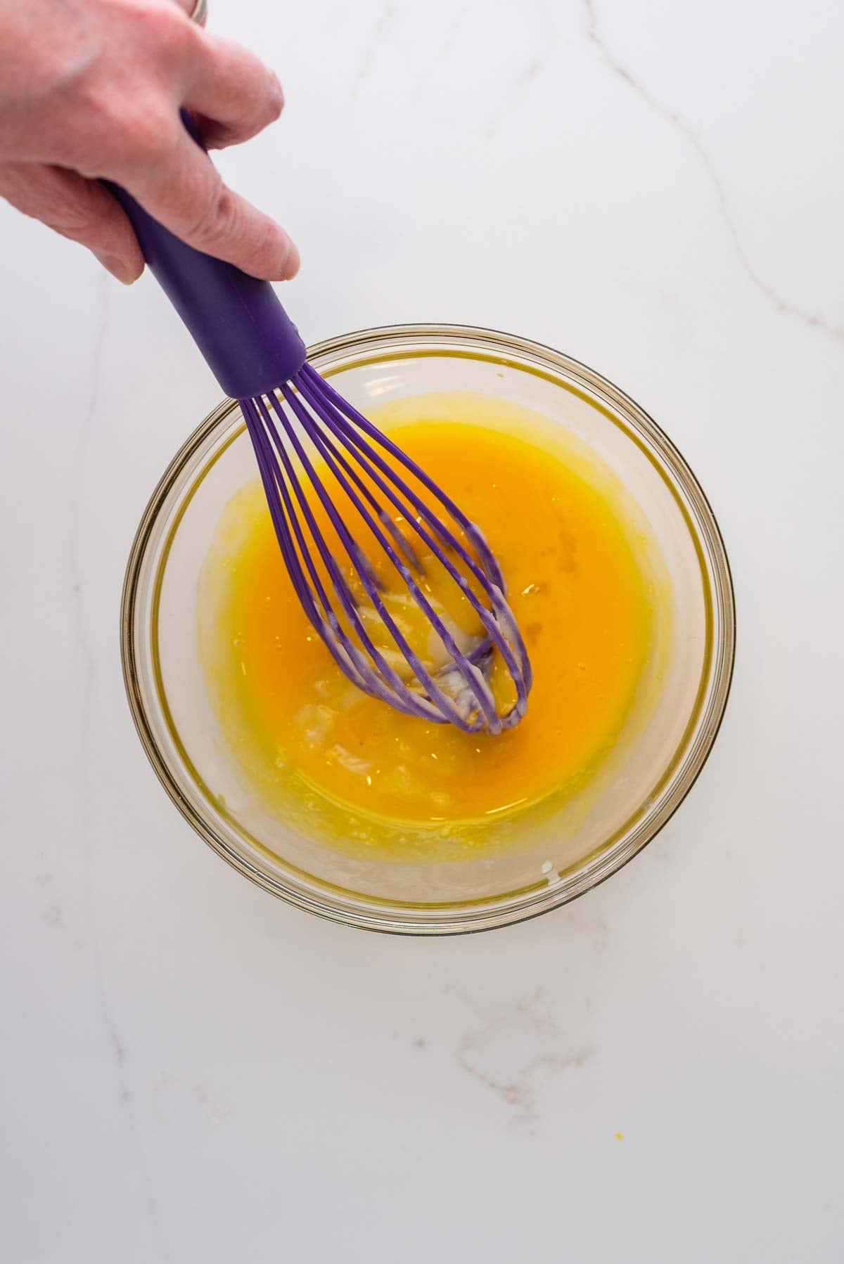 Tempering egg yolks with lemon pie filling mixture in a small glass bowl and purple whisk on a white marbled background.