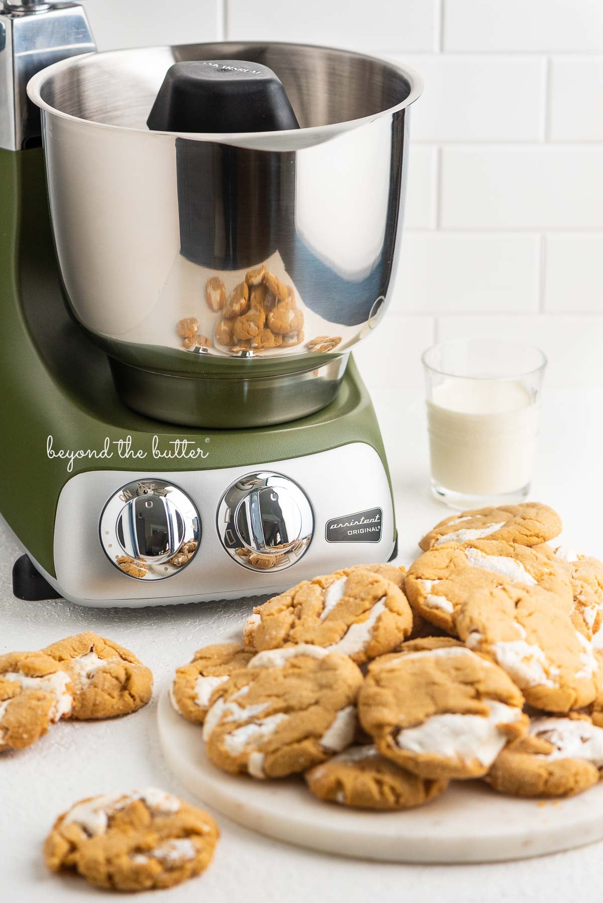 Olive green Ankarsrum stand mixer with fluffernutter cookies and glass of milk on white background.