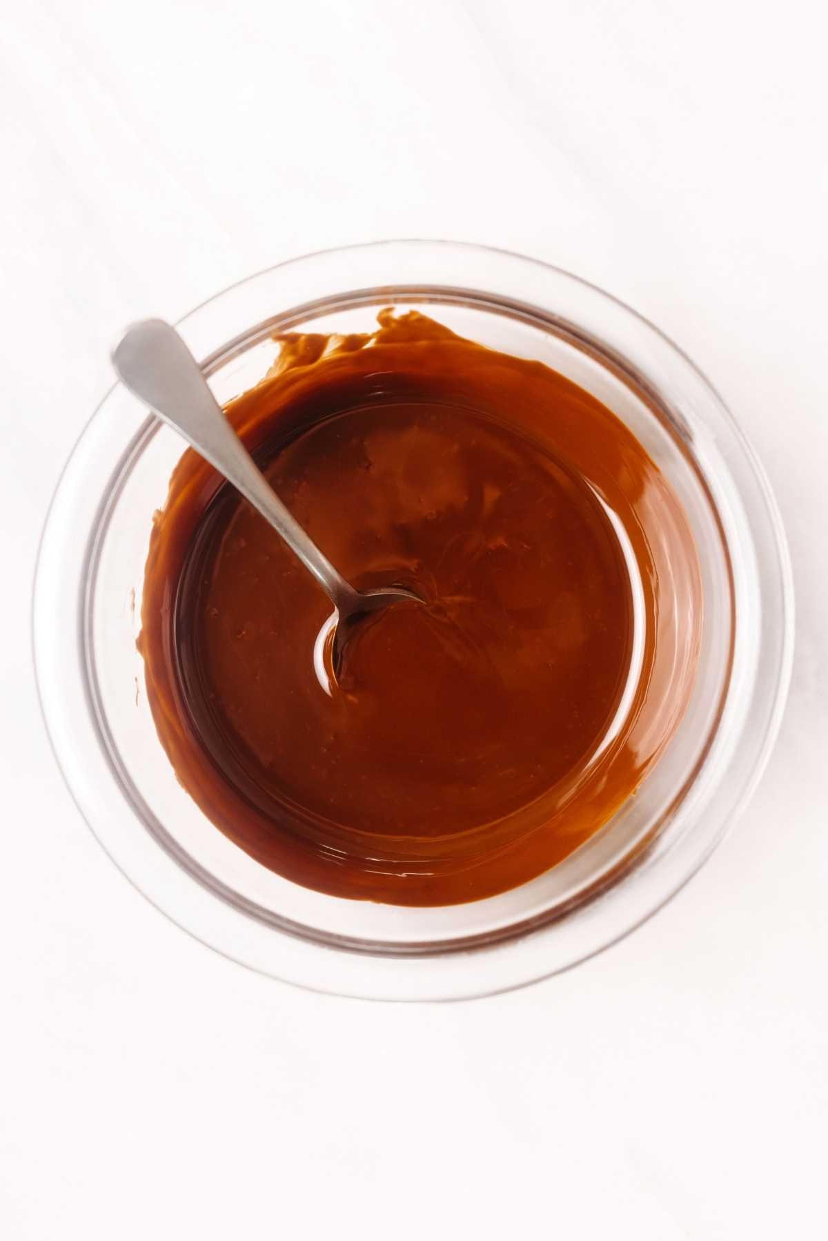 Melted dark chocolate in small glass bowl with a spoon resting in the chocolate on a white textured background.