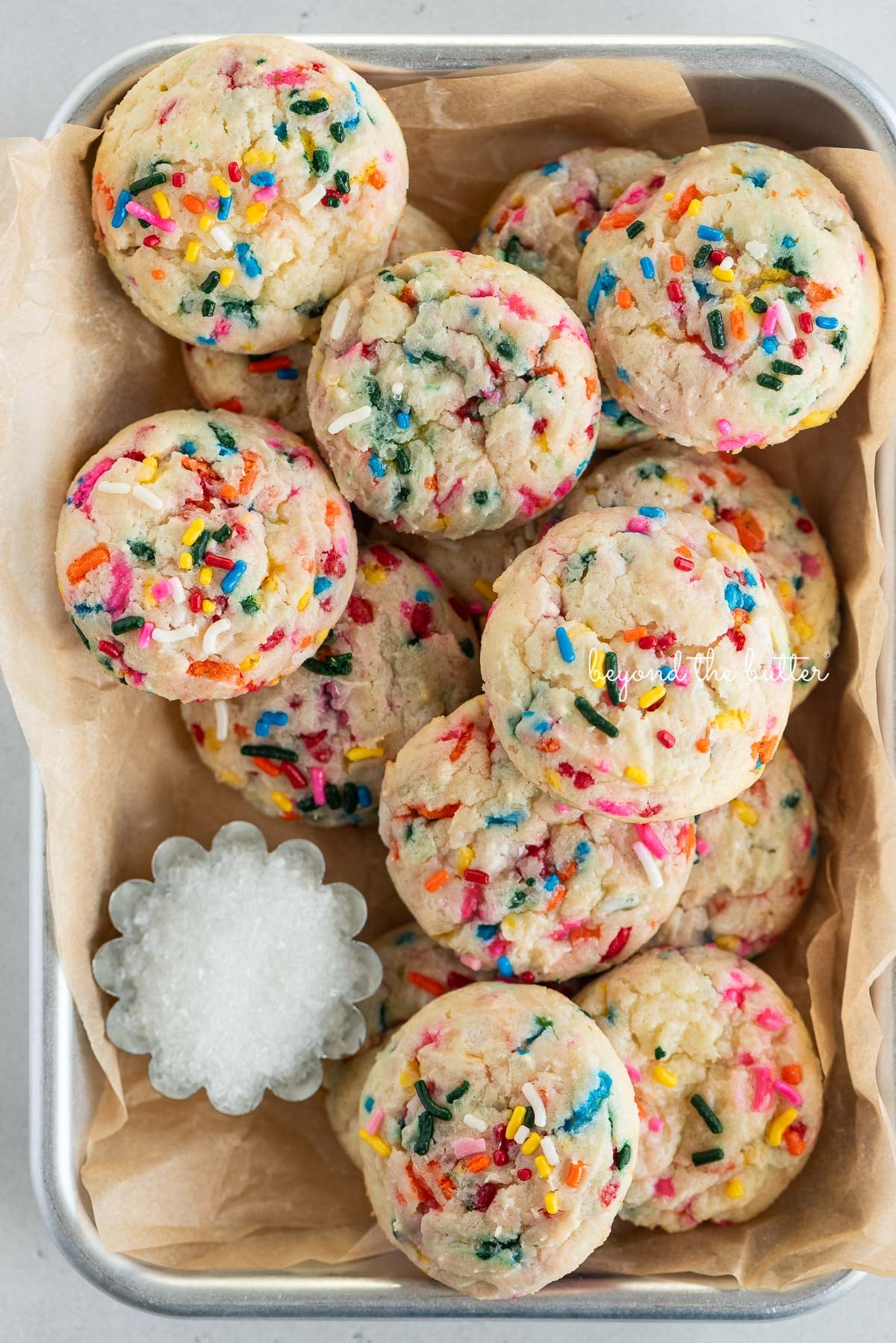 Parchment paper lined small baking sheet filled with funfetti butter cookies and a small scalloped bowl of white sparkling sugar.