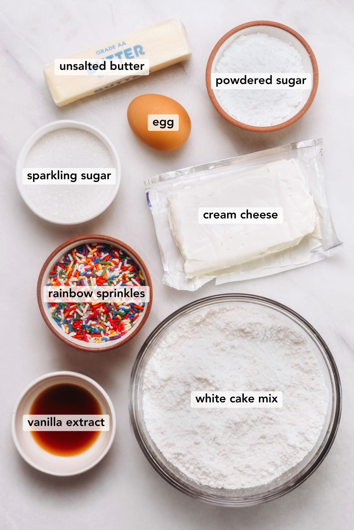 Ingredients in various size bowls labeled as all unsalted butter, powdered sugar, egg, sparkling sugar, cream cheese, rainbow sprinkles, white cake mix, and vanilla extract on a white marbled background.