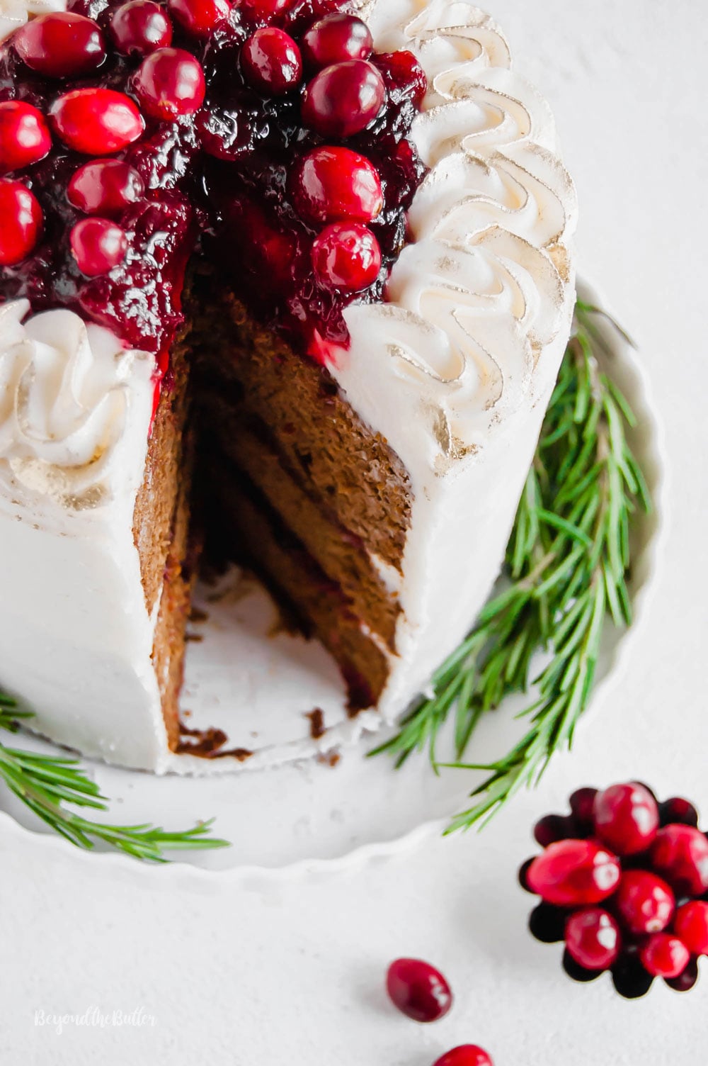 Gingerbread Cake | Angled photo of Gingerbread Cake with Maple Buttercream Frosting and Cranberry Compote with a slice taken out of it and a little bowl of cranberries below it | Image Credit: Luyiz, LLC