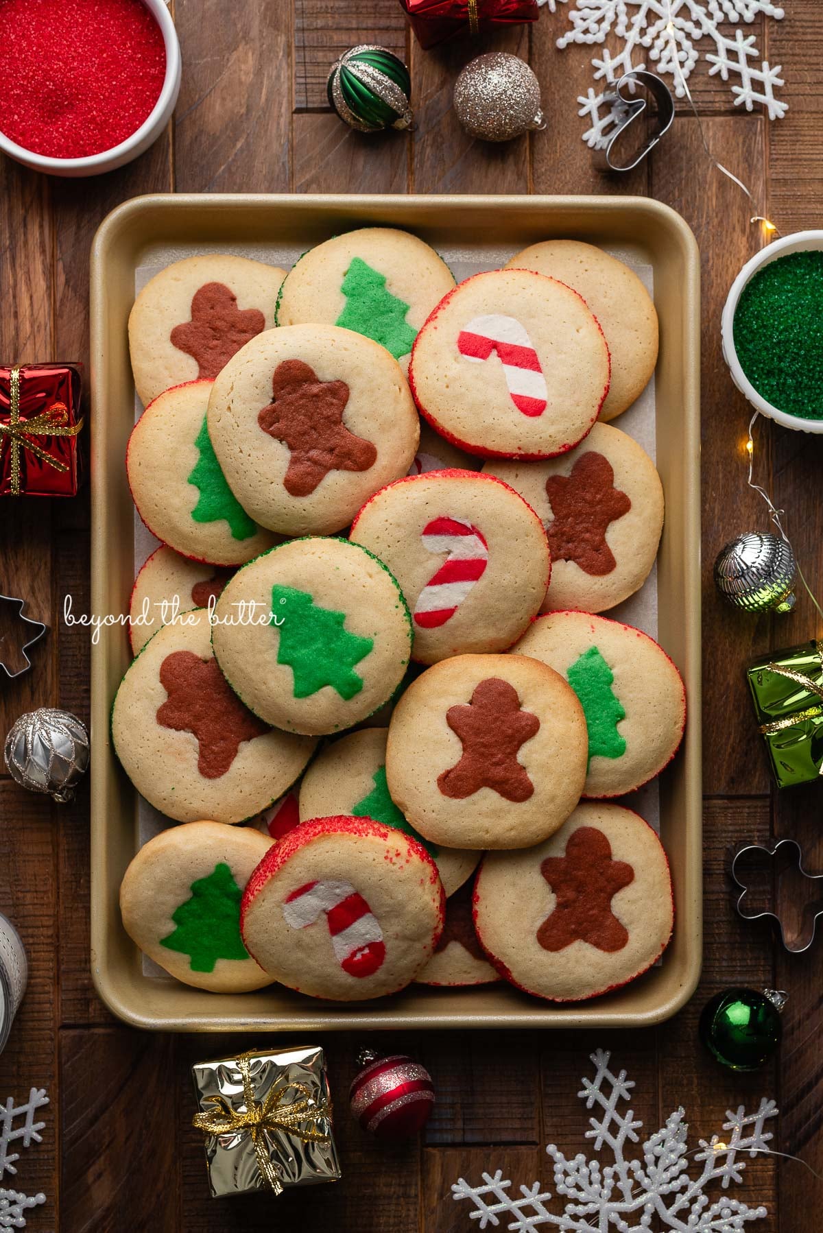 Slice and bake holiday cookies on a gold colored cookie sheet surrounded by ornaments, lights, cookie cutters, and sparkling sugars on a dark wooden background.