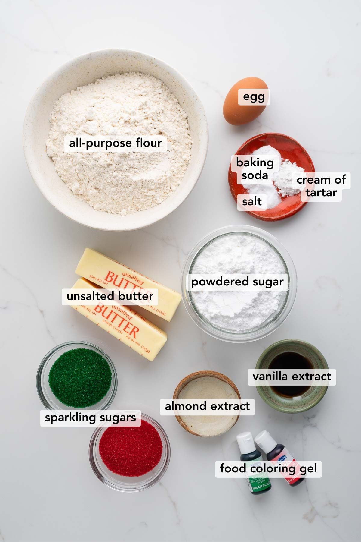 Ingredients in various size bowls and plates labeled as all purpose flour, egg, baking soda, cream of tartar, salt, unsalted butter, powdered sugar, sparkling sugars, almond extract, vanilla extract, and food coloring gel on a white marbled background.