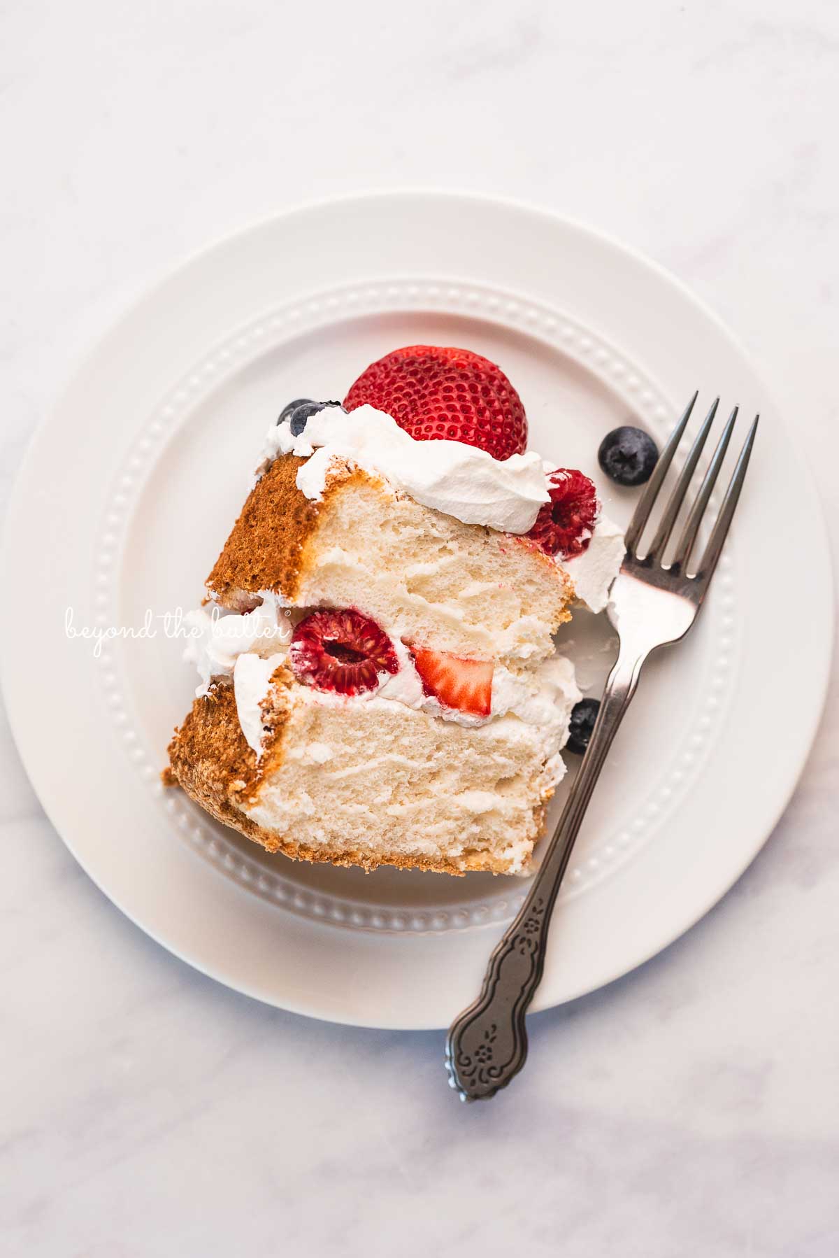 Slice of homemade angel food cake with berries on white dessert plate and white marbled background | © Luyiz®