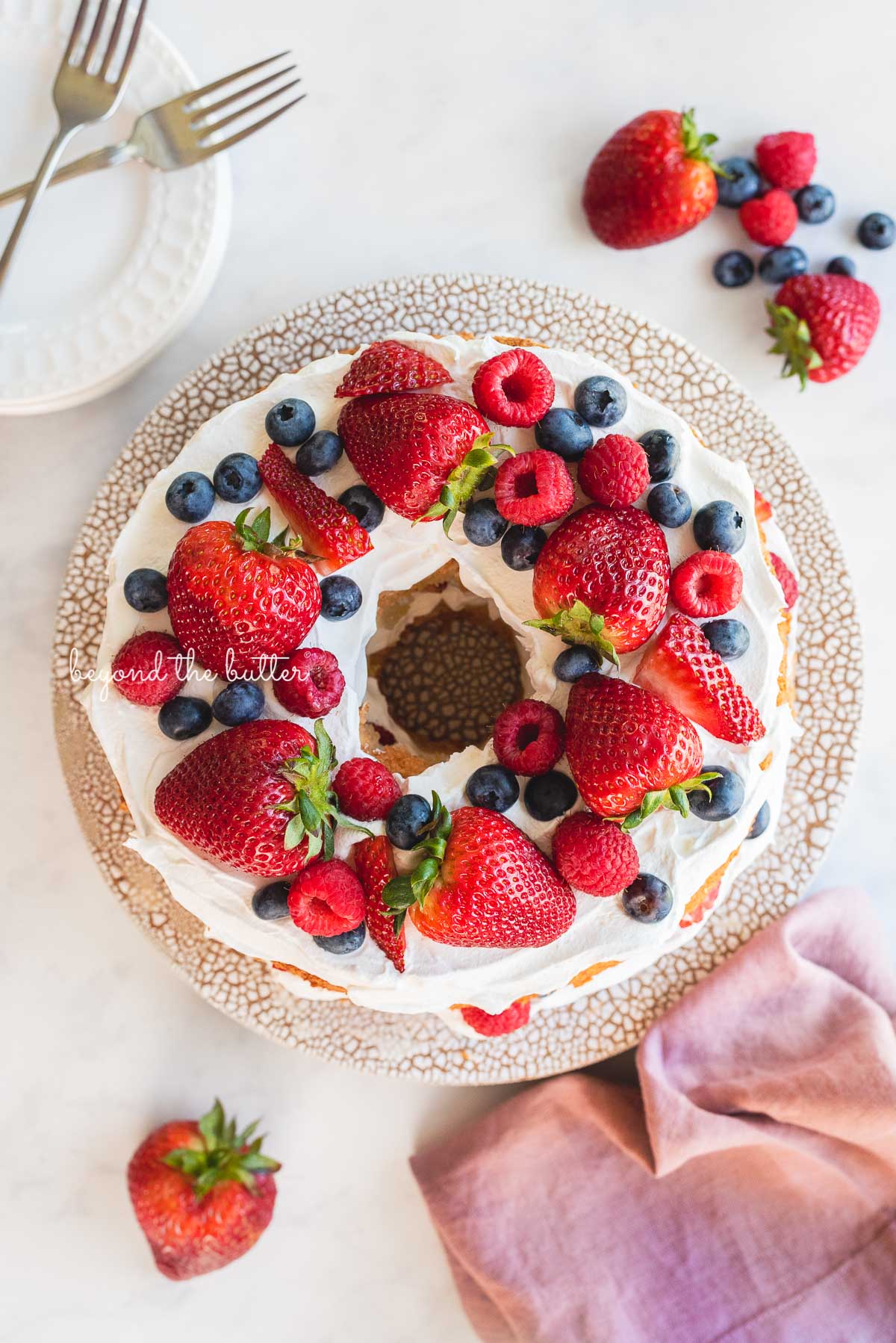 Homemade angel food cake on ceramic cake stand surrounded by dessert plates, forks, berries, and mauve colored cloth napkin on white marbled background | © Luyiz®