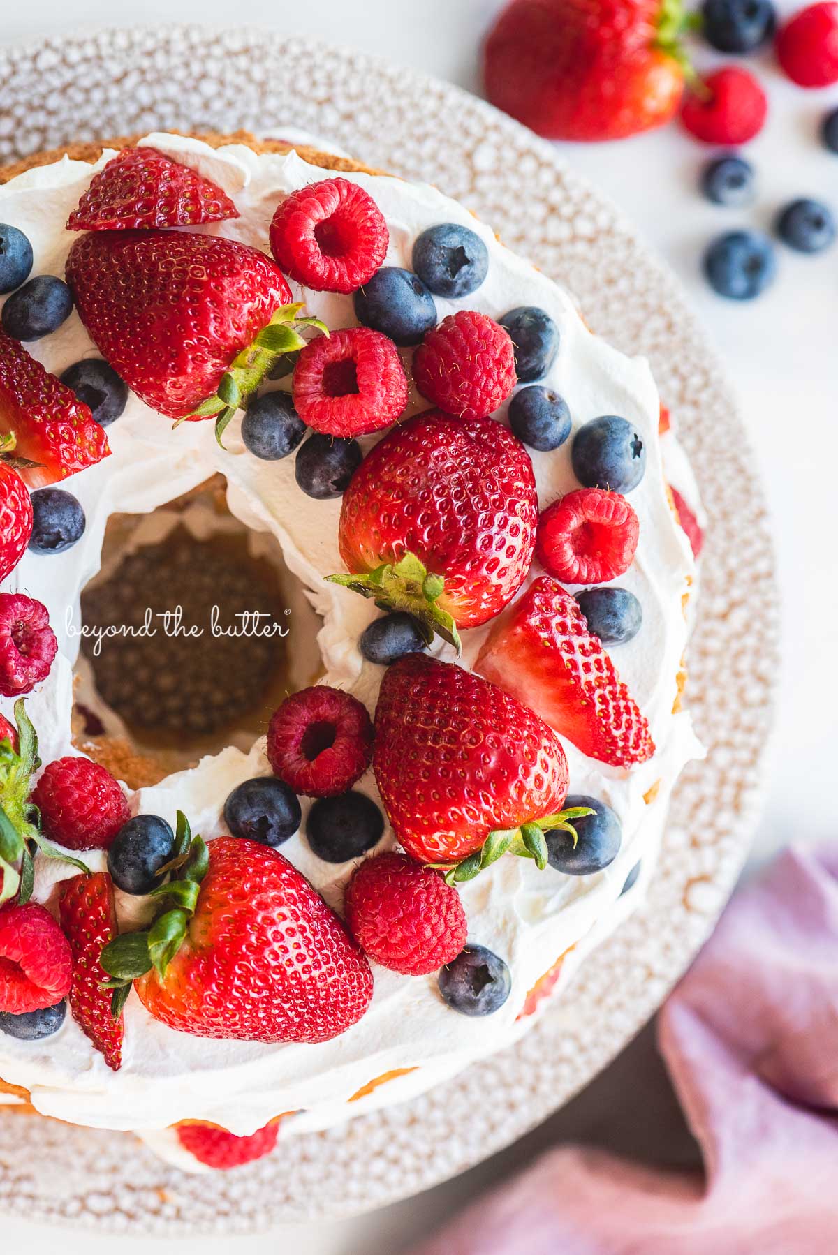 Homemade angel food cake with berries on ceramic cake stand surrounded by berries, and mauve colored cloth napkin on white marbled background | © Luyiz®