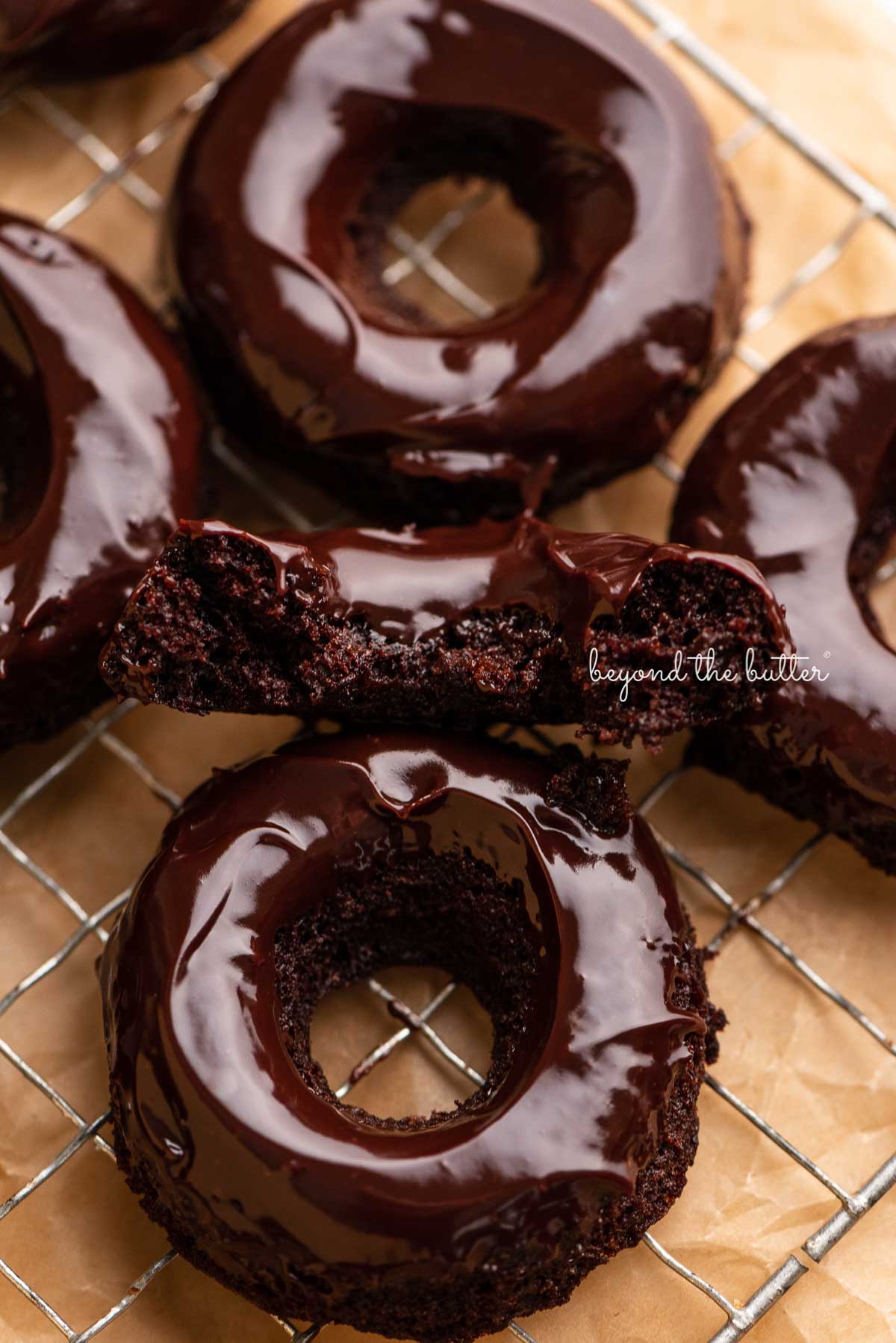 Half of a homemade baked chocolate donut topped with chocolate ganache on a wire cooling rack with more donuts | © Luyiz®