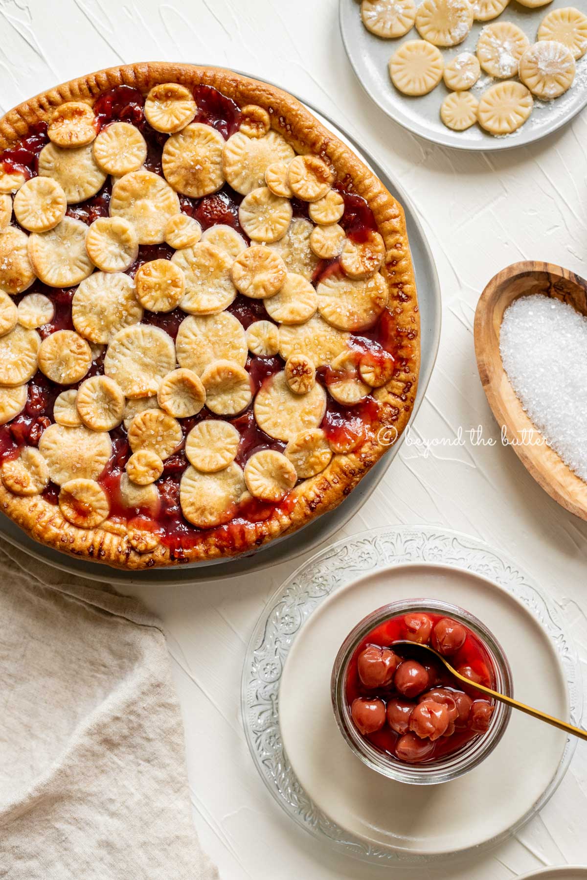 Overhead image of cherry pie surrounded by plates, jar of cherries, sprakling sugar, and pie crust toppers | All Images © Luyiz™