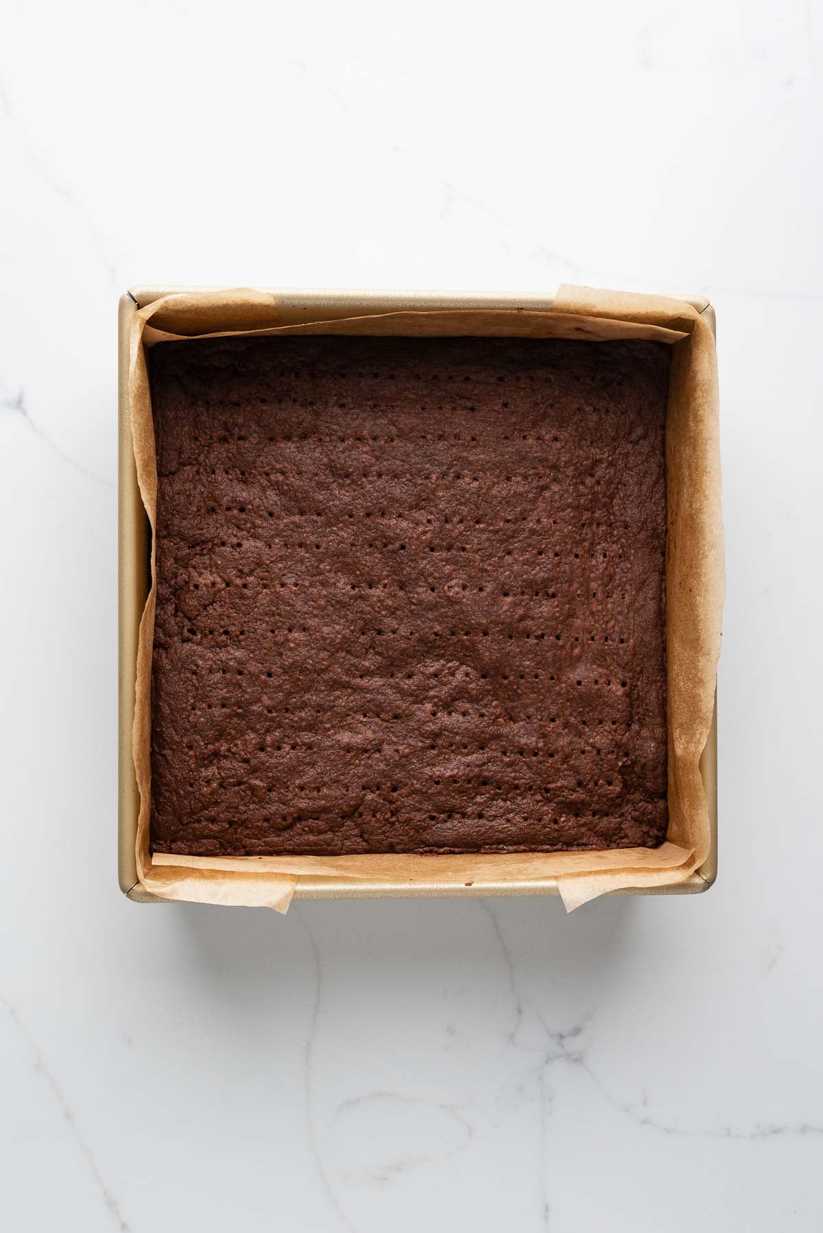 Baked chocolate shortbread biscuits in a parchment paper lined metal baking tin on a white marbled surface.