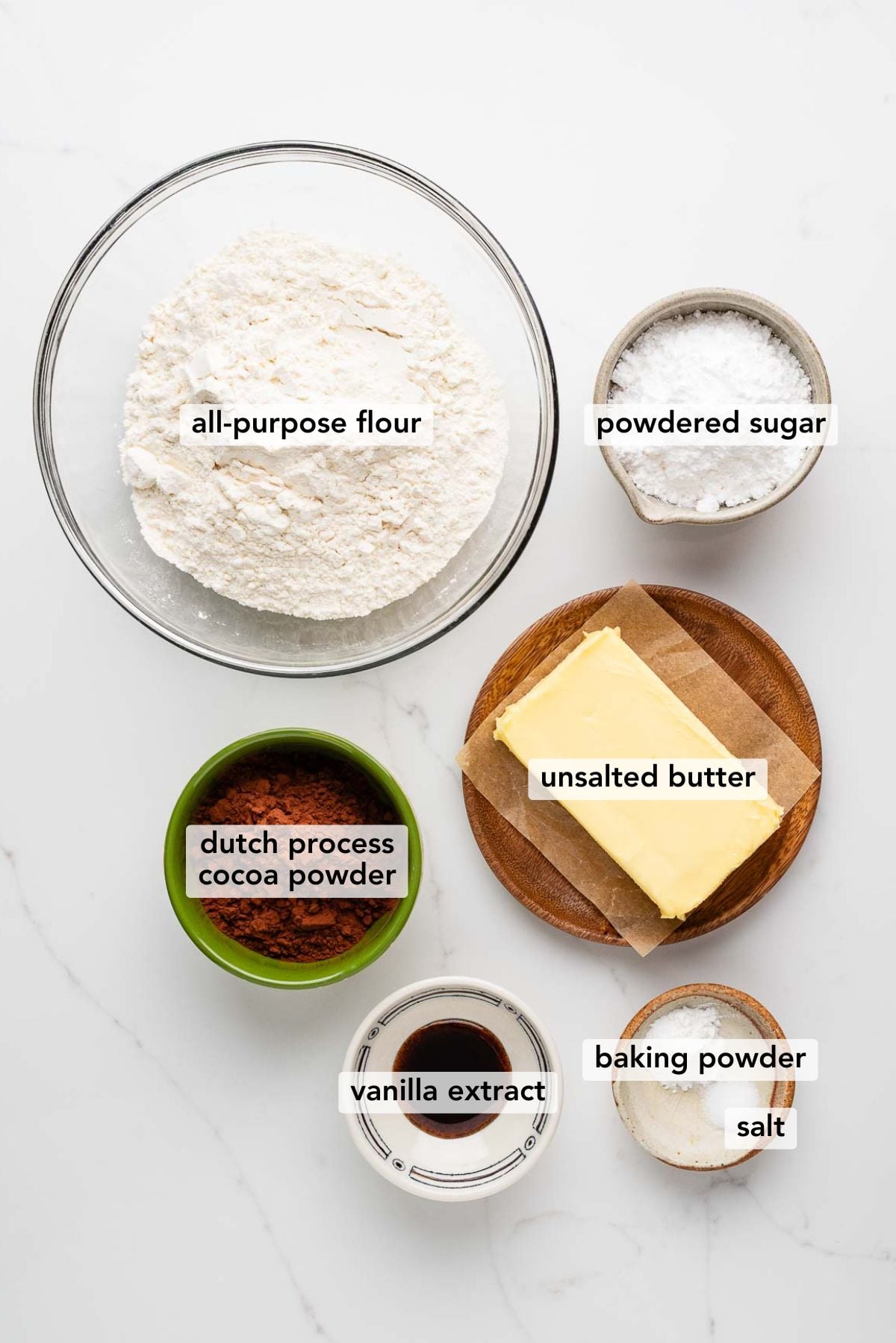 Ingredients in various size bowls labeled as all purpose flour, powdered sugar, dutch process cocoa powder, unsalted butter, vanilla extract, baking powder, and salt on a white marbled background.
