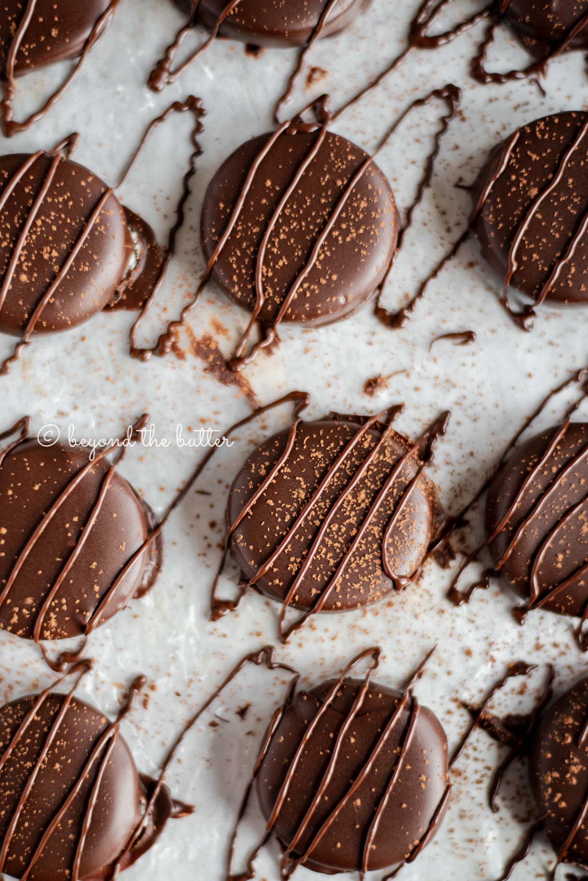 Round chocolate dipped peppermint patties drizzled with chocolate and dusted with cocoa powder on wax paper lined baking sheet.