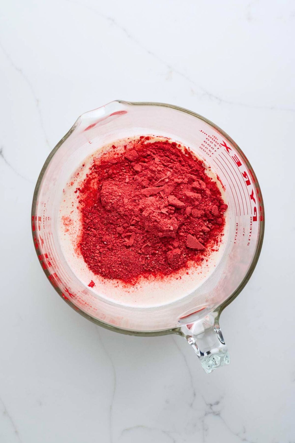 Strawberry ice cream ingredients added to a large glass mixing bowl on a white marbled background.