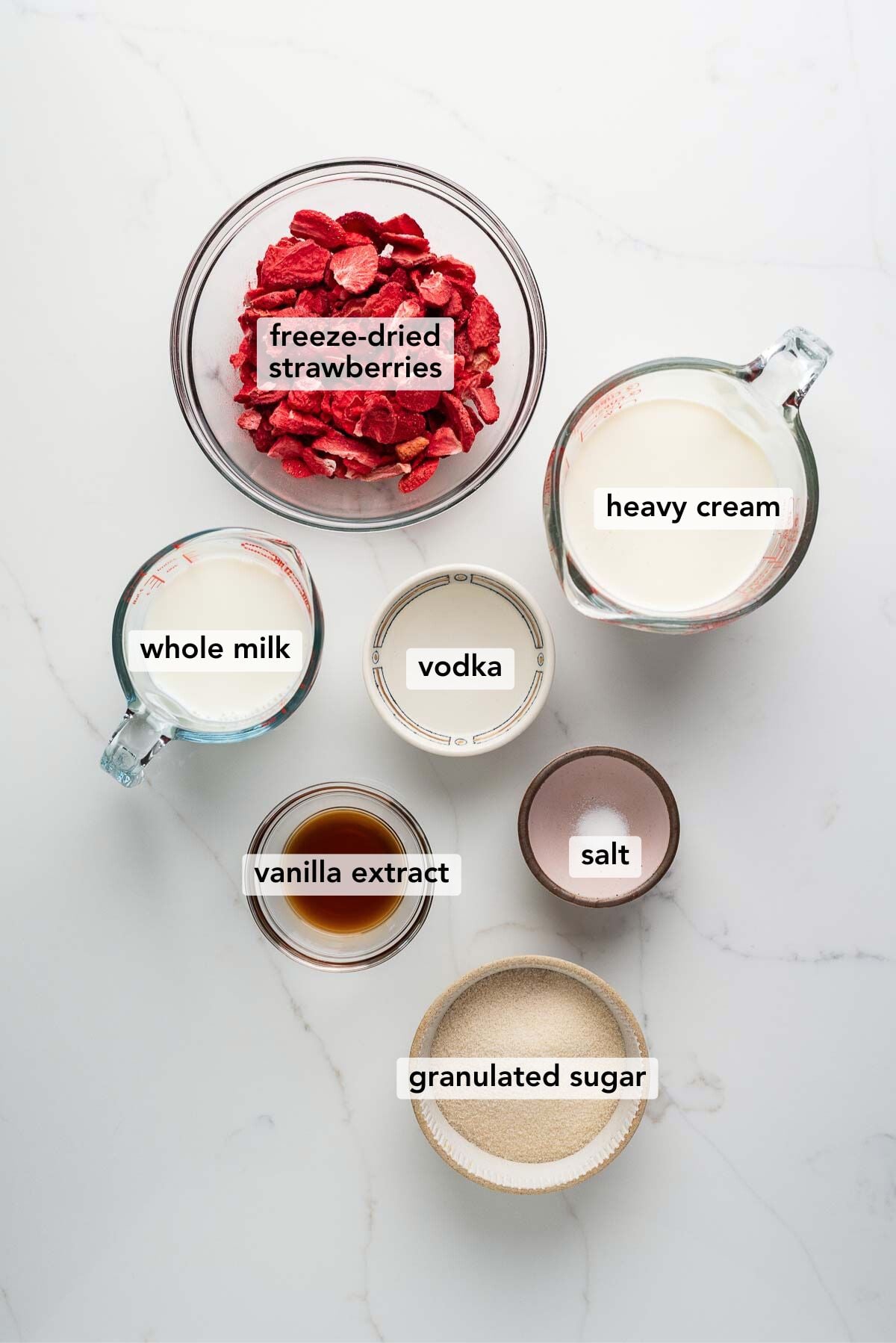 Ingredients in various size plates and bowls labeled as freeze-dried strawberries, heavy cream, whole milk, vodka, vanilla extract, salt, and granulated sugar on a white marbled background.