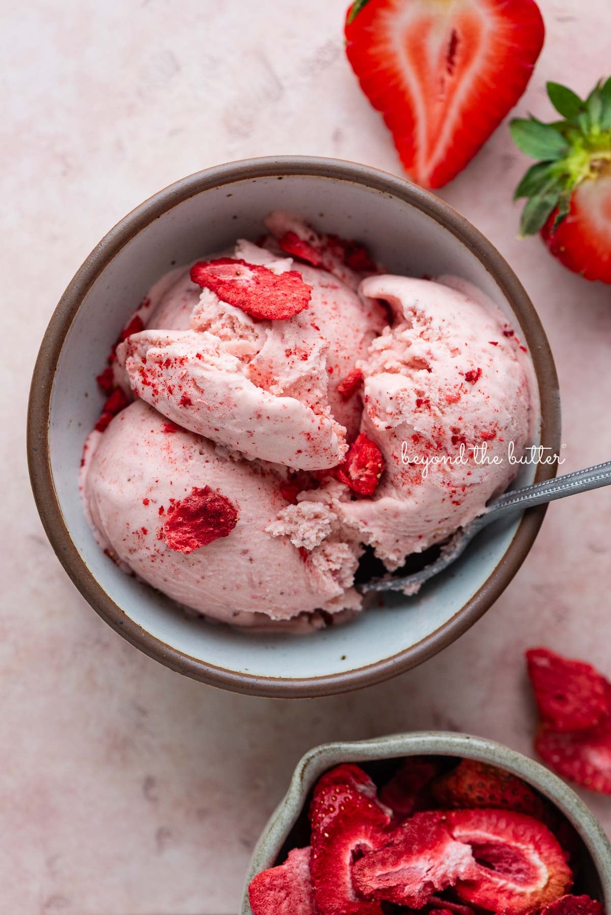Small while ceramix bowl with homemade strawberry ice cream topped with bits of freeze-dried strawberries with a small bowl of freeze-dried strawberries and fresh strawberries surrounding it on a pink tiled background.
