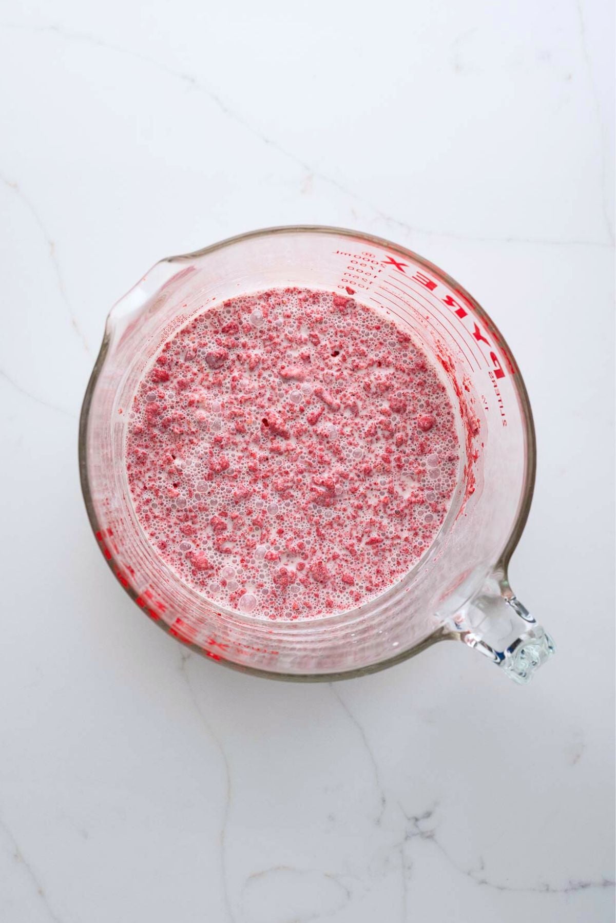 Strawberry ice cream base mixed all together in a large glass mixing bowl on a white marbled background.