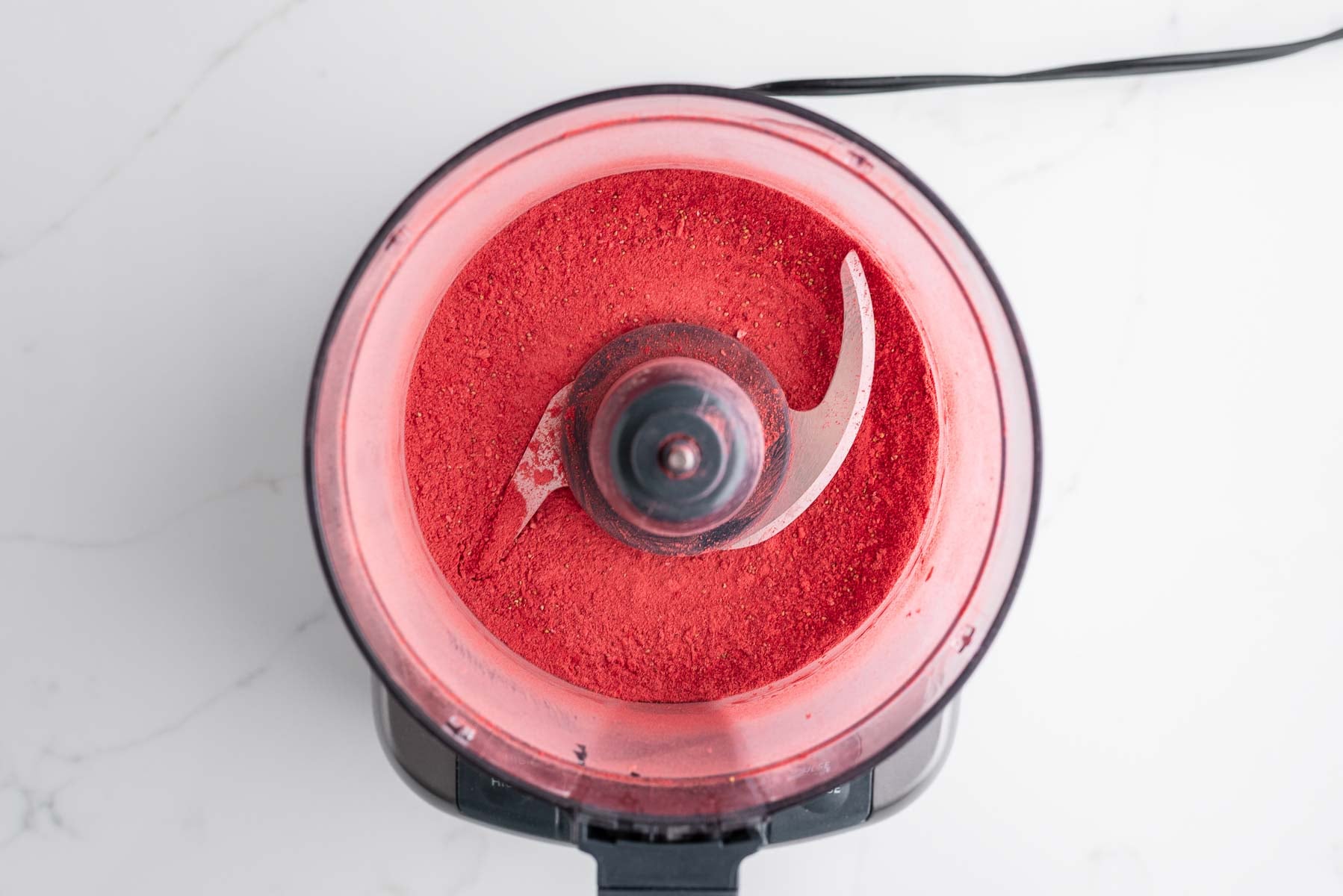 Processed freeze-dried strawberries in a medium-sized food processor on a white marbled background.