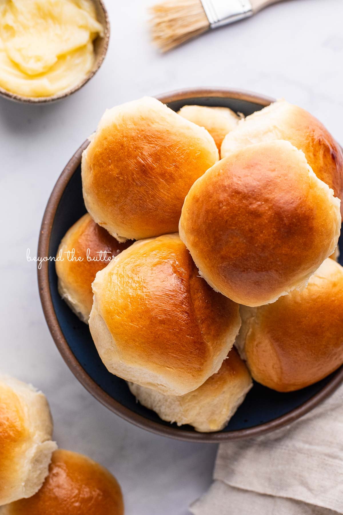 Blue ceramic bowl filled with homemade honey butter dinner rolls with a small bowl of honey butter and brush next to it on a white marbled background | © Luyiz®