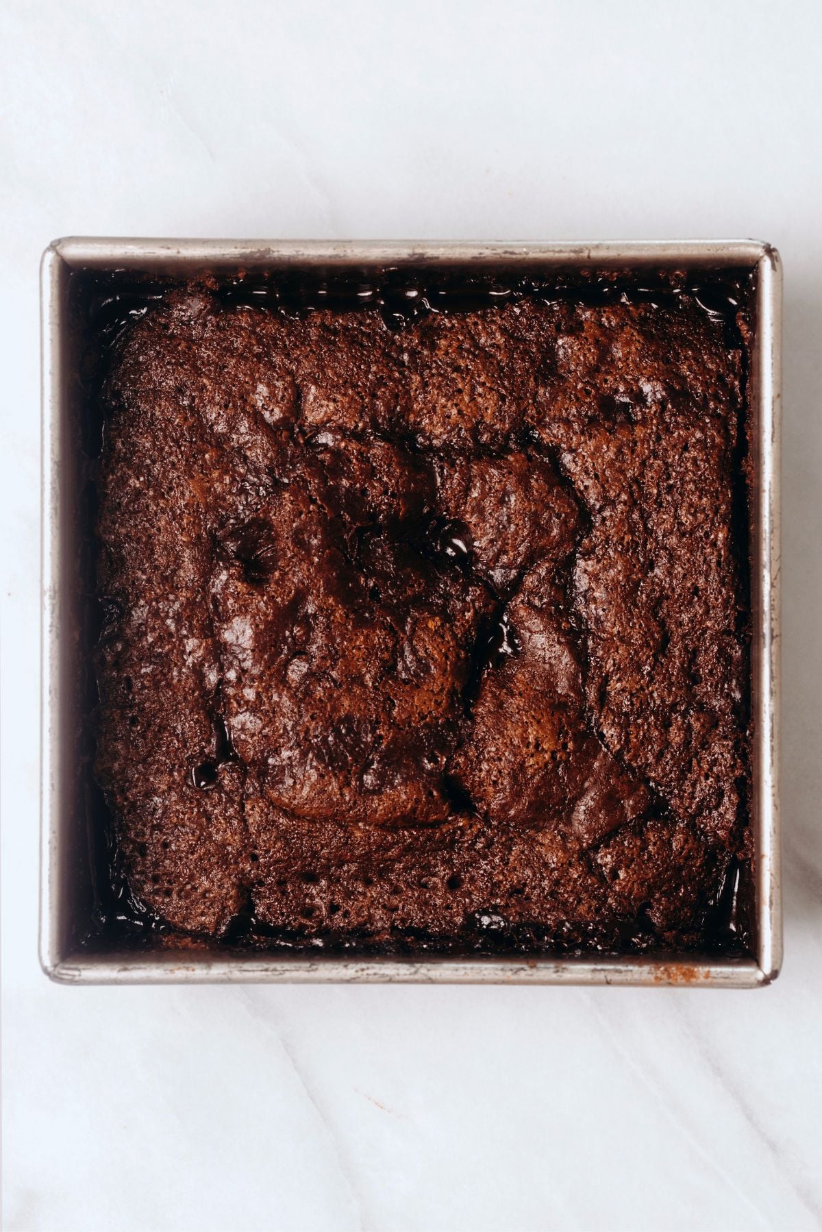 Hot fudge chocolate pudding cake baked in a square metal baking tin on a white marbled background.