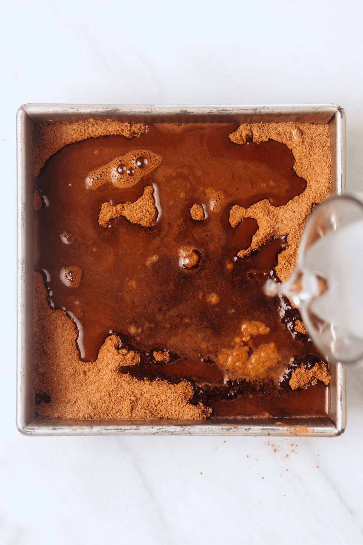 Pouring boiling water over hot fudge pudding cake batter in a metal baking tin on a white marbled background.