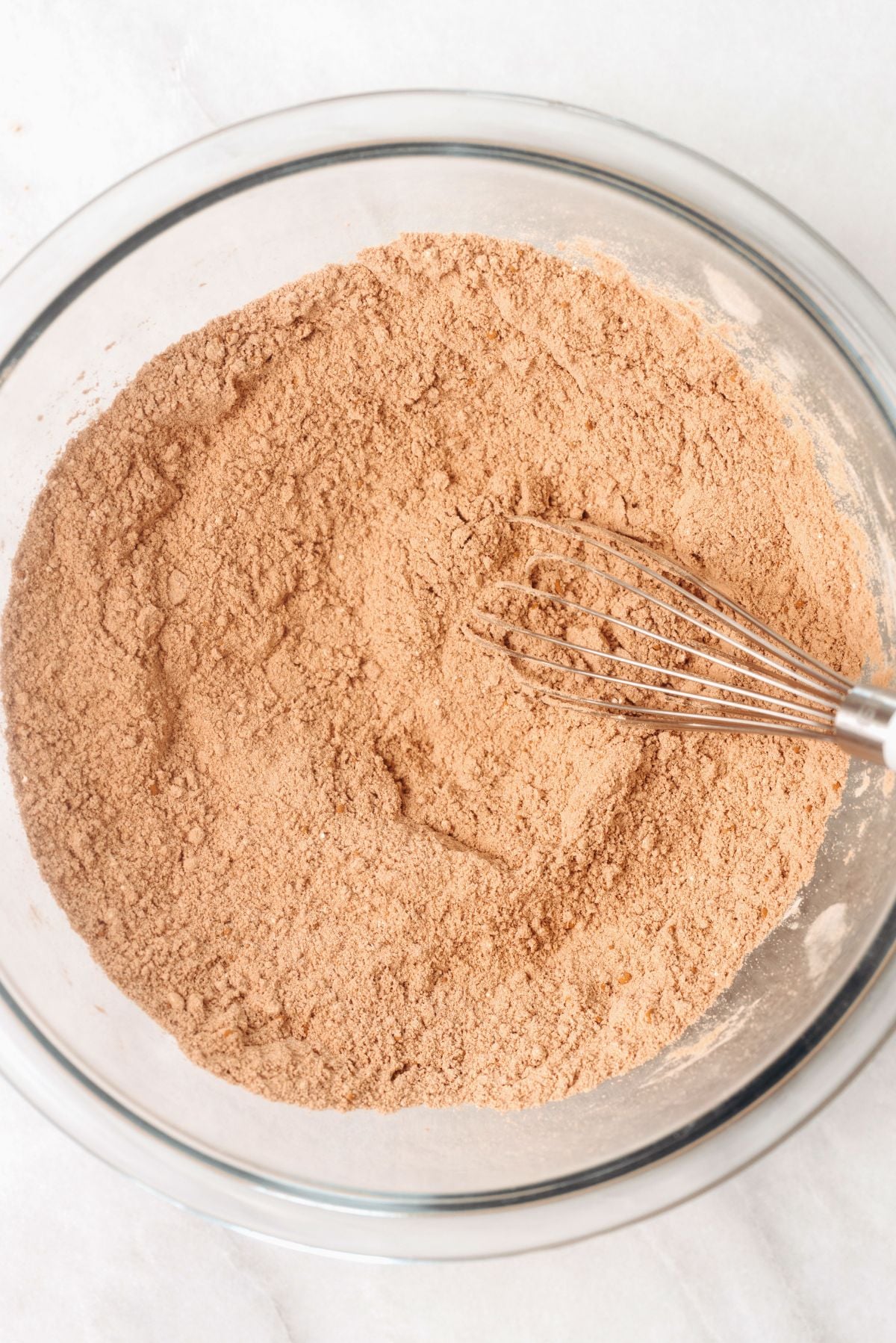 Sugar, flour, cocoa, baking powder, and salt whisked together in a large glass mixing bowl on a white marbled background.
