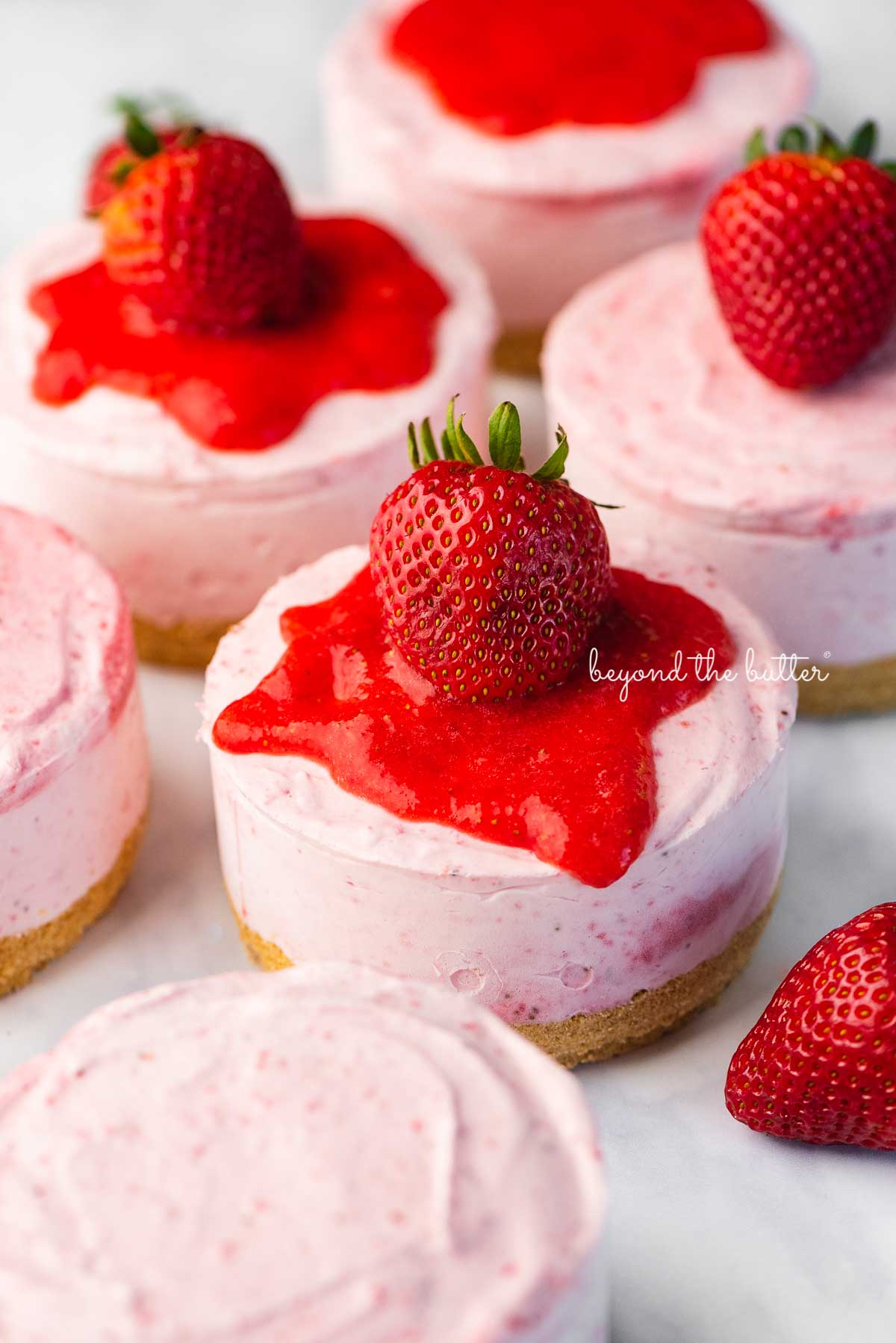 Individual no-bake strawberry cheesecakes topped with strawberry glaze and a fresh strawberry on a parchment paper-lined baking sheet | © Luyiz®