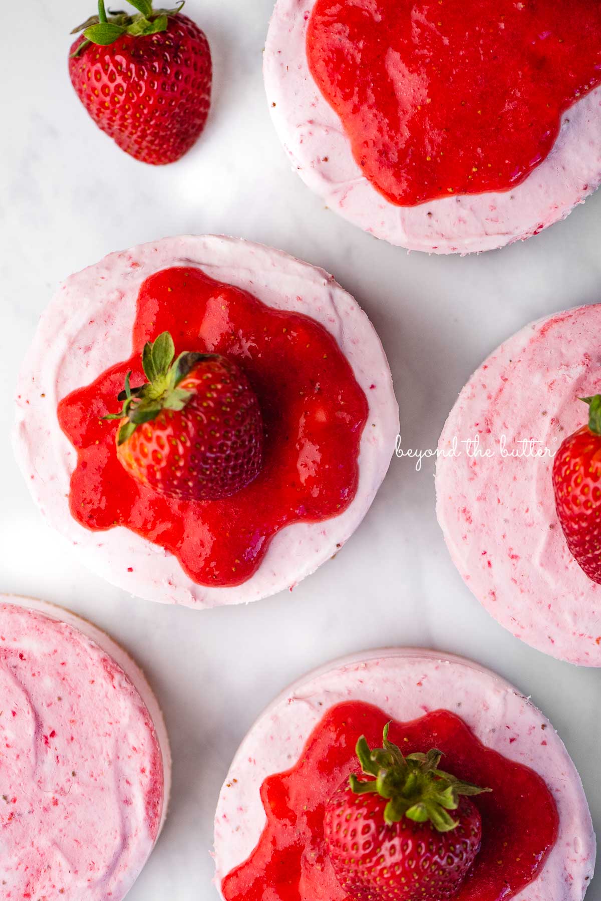 Small 4-inch no-bake strawberry cheesecakes topped with strawberry glaze and a fresh strawberry on a parchment paper-lined baking sheet | © Luyiz®