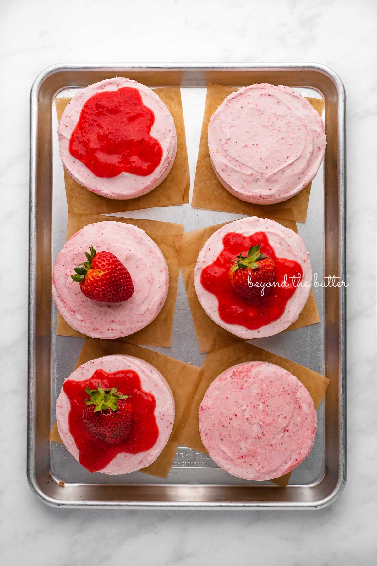 Individual no-bake strawberry cheesecakes topped with strawberry glaze and a fresh strawberry on a parchment paper-lined baking sheet | © Luyiz®