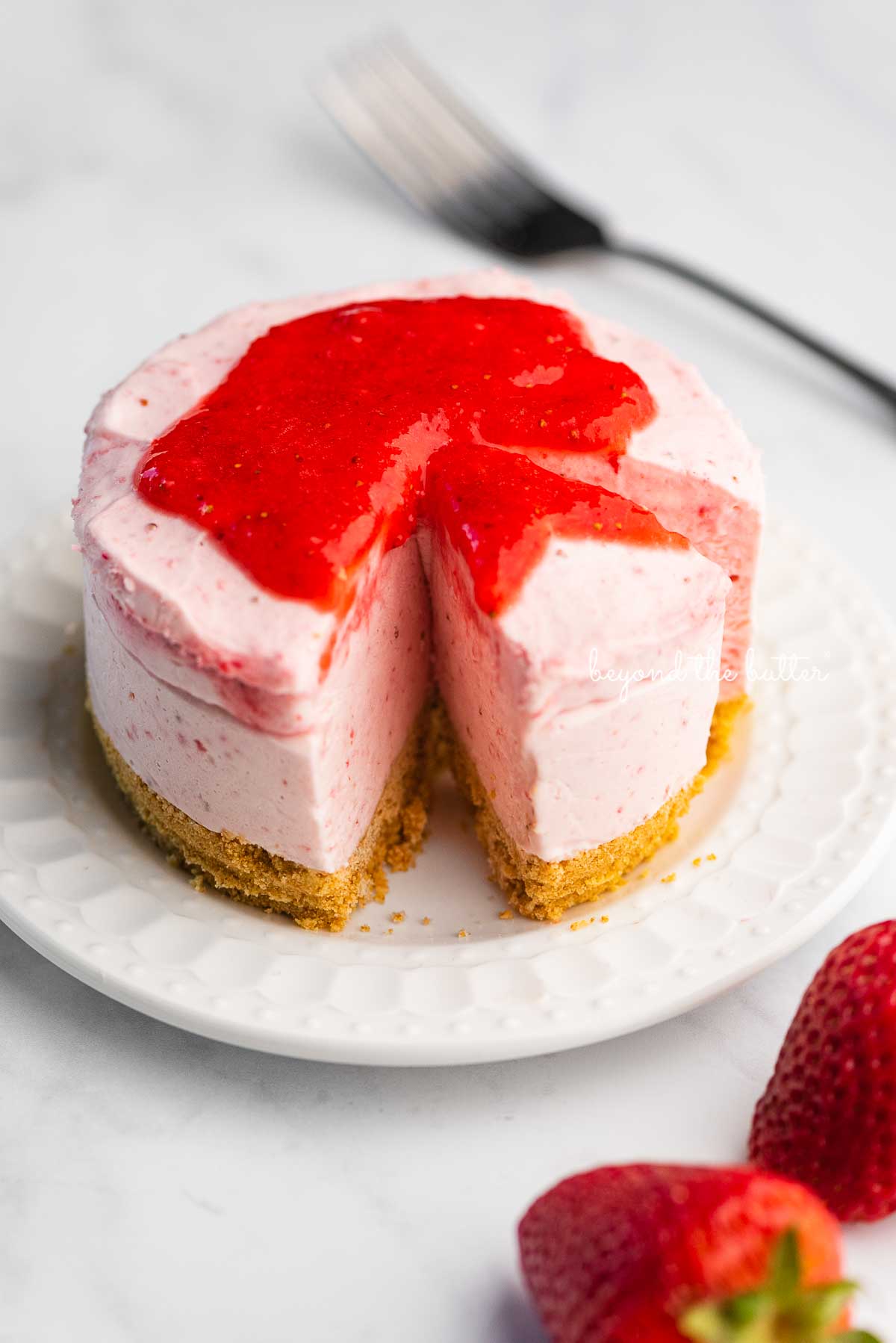 Small 4-inch no-bake strawberry cheesecake on a white dessert plate with a fork and fresh strawberries nearby on a marbled background | © Luyiz®