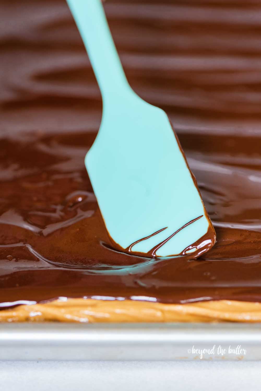 Angled view of blue spatula spreading chocolate over the top of peanut butter tandy kakes | All Images © Luyiz™