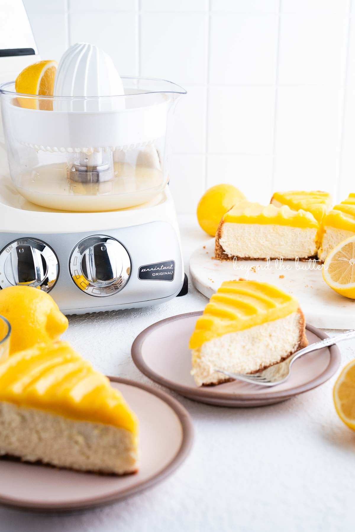 Slices of lemon cheesecake topped with lemon curd on pink dessert plates with cream colored Ankarsrum mixer on white background.