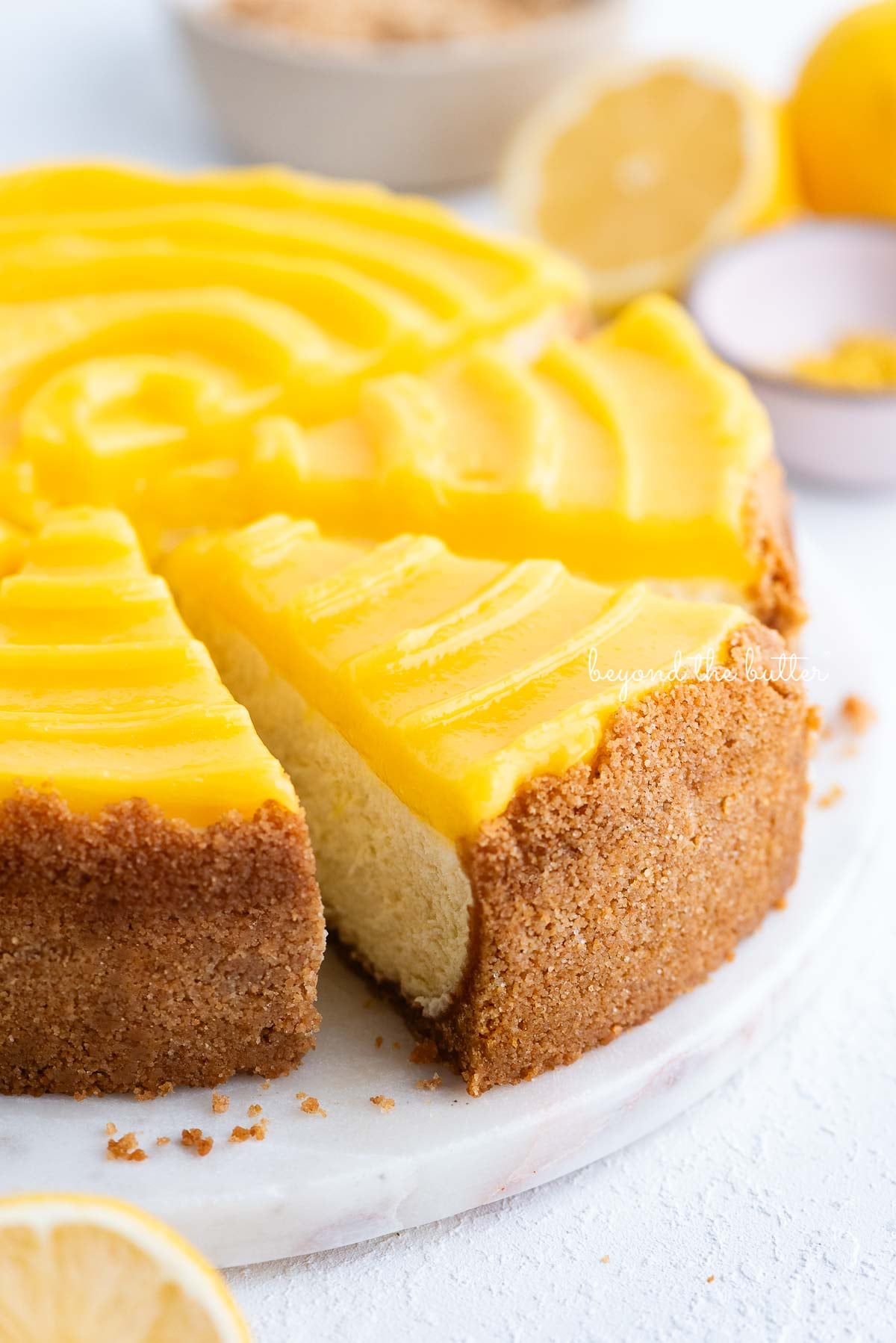Slice of lemon cheesecake topped with lemon curd being removed from the cake plate with cake server on a white background.